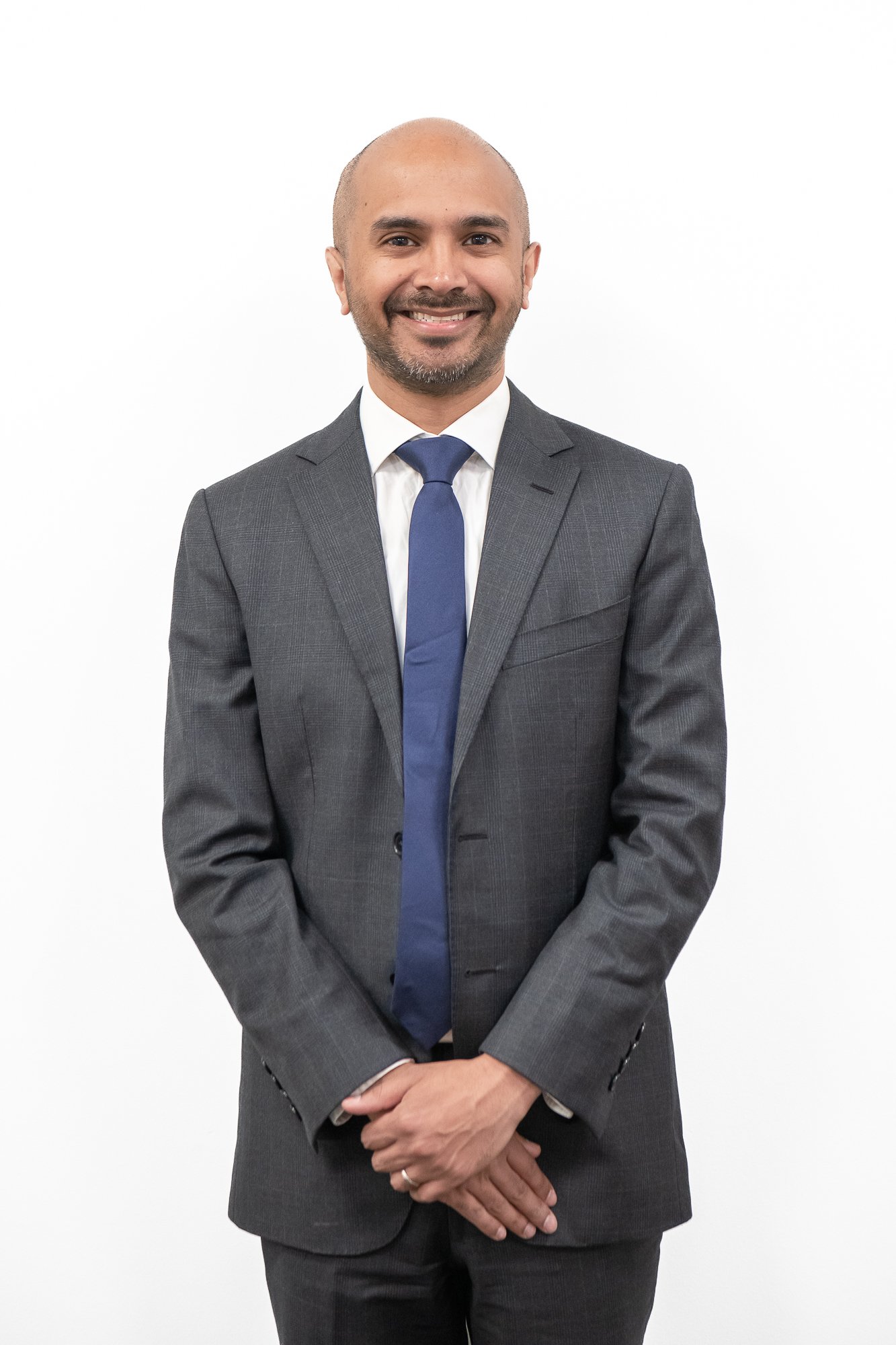 A man wearing a gray suit with a white shirt and blue tie, smiling and standing against a white background.
