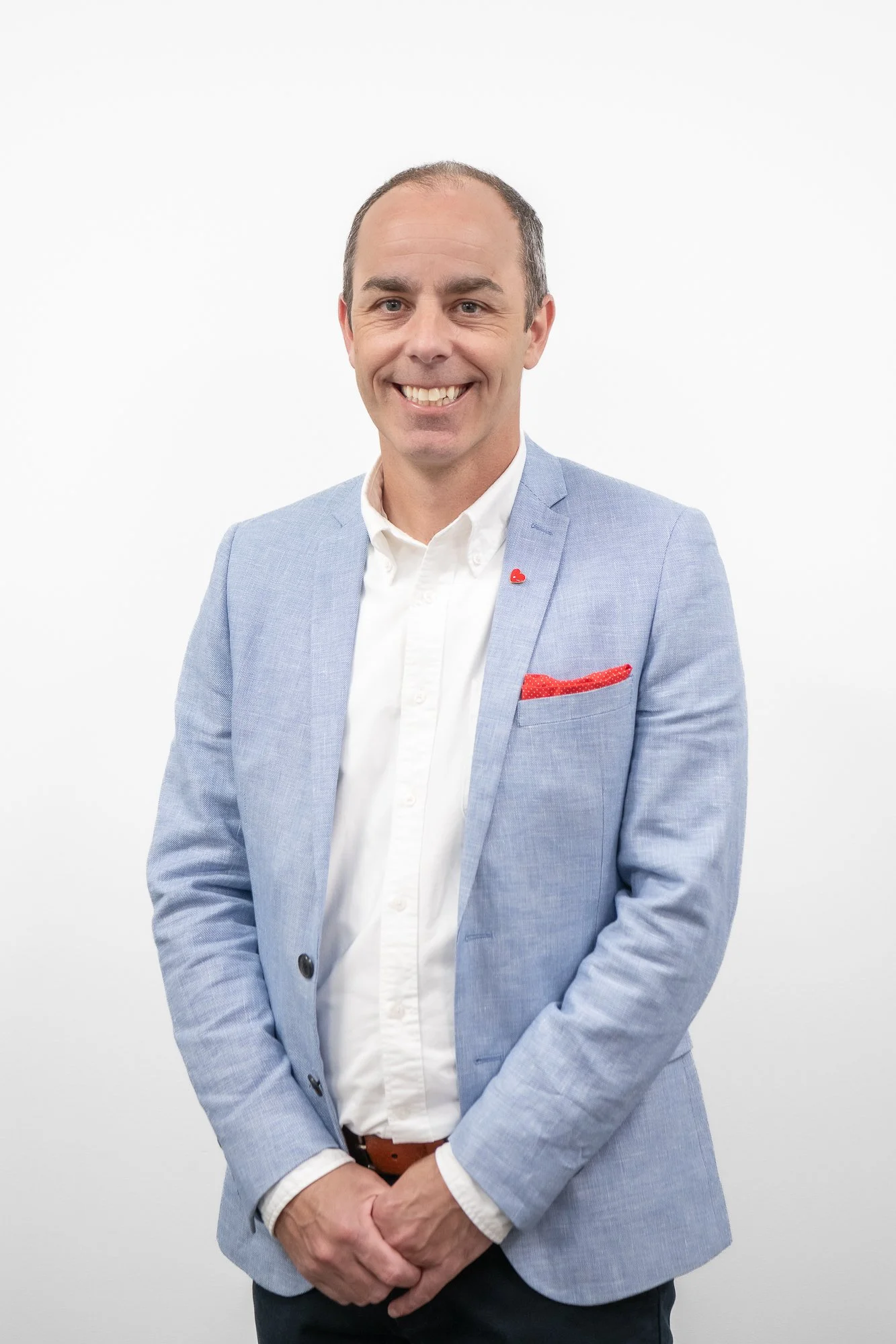 A smiling man with short hair, wearing a light blue blazer, white shirt, and a red pocket square, standing against a plain white background.