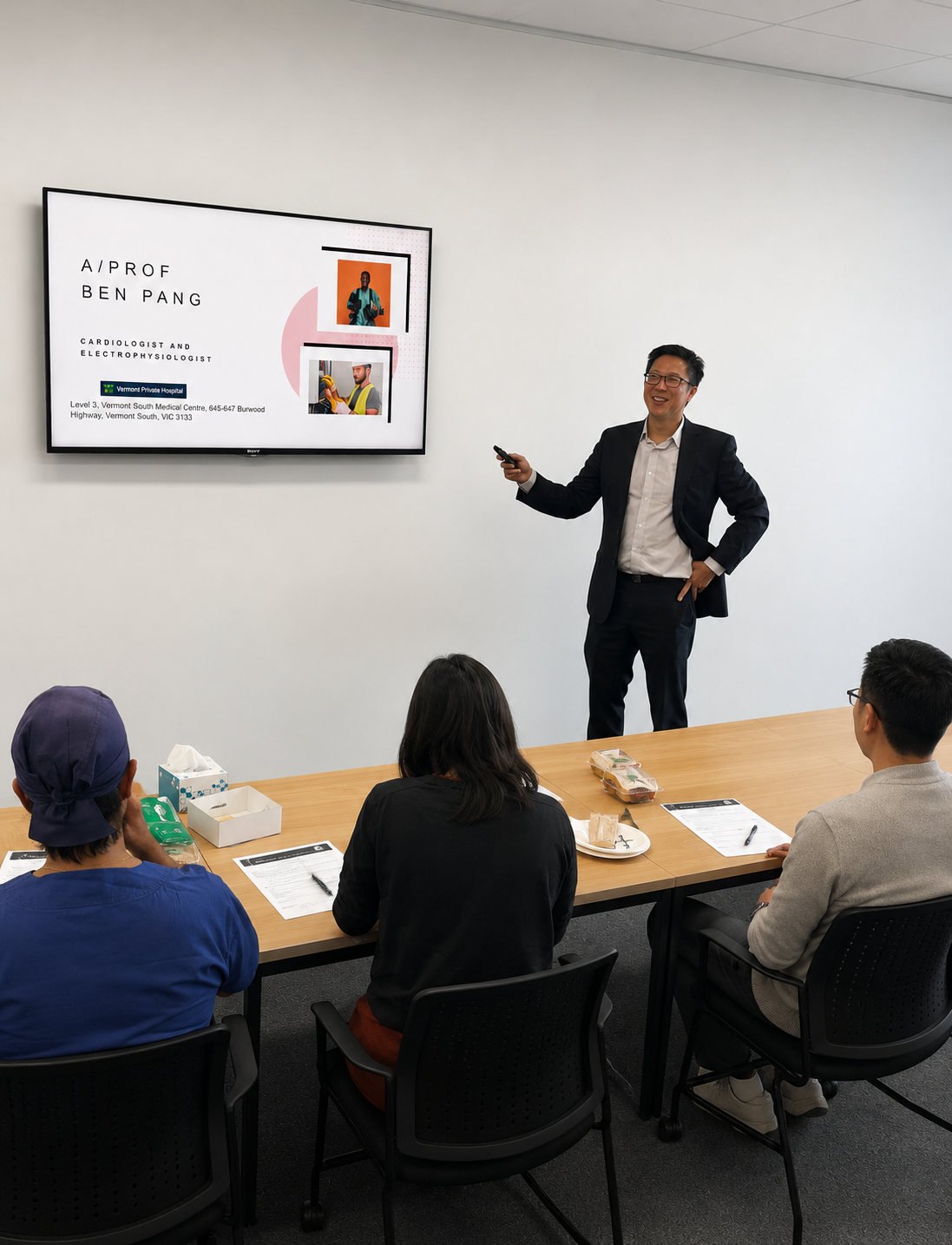 A man in a suit giving a presentation to three seated attendees in a conference room, with a large screen displaying his name and credentials.