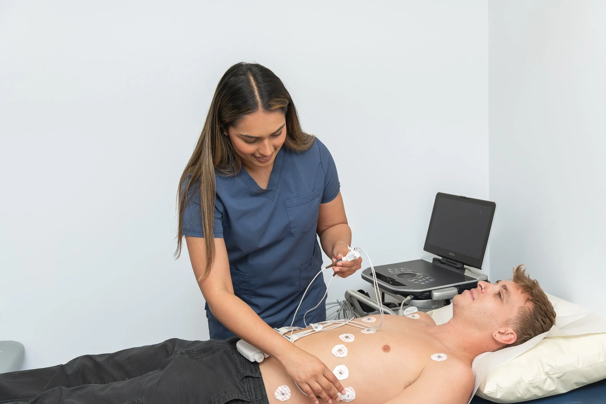 A healthcare professional performing an electrocardiogram (ECG) on a male patient lying in a hospital bed. The patient has electrical sensors attached to his chest and a monitor next to him.