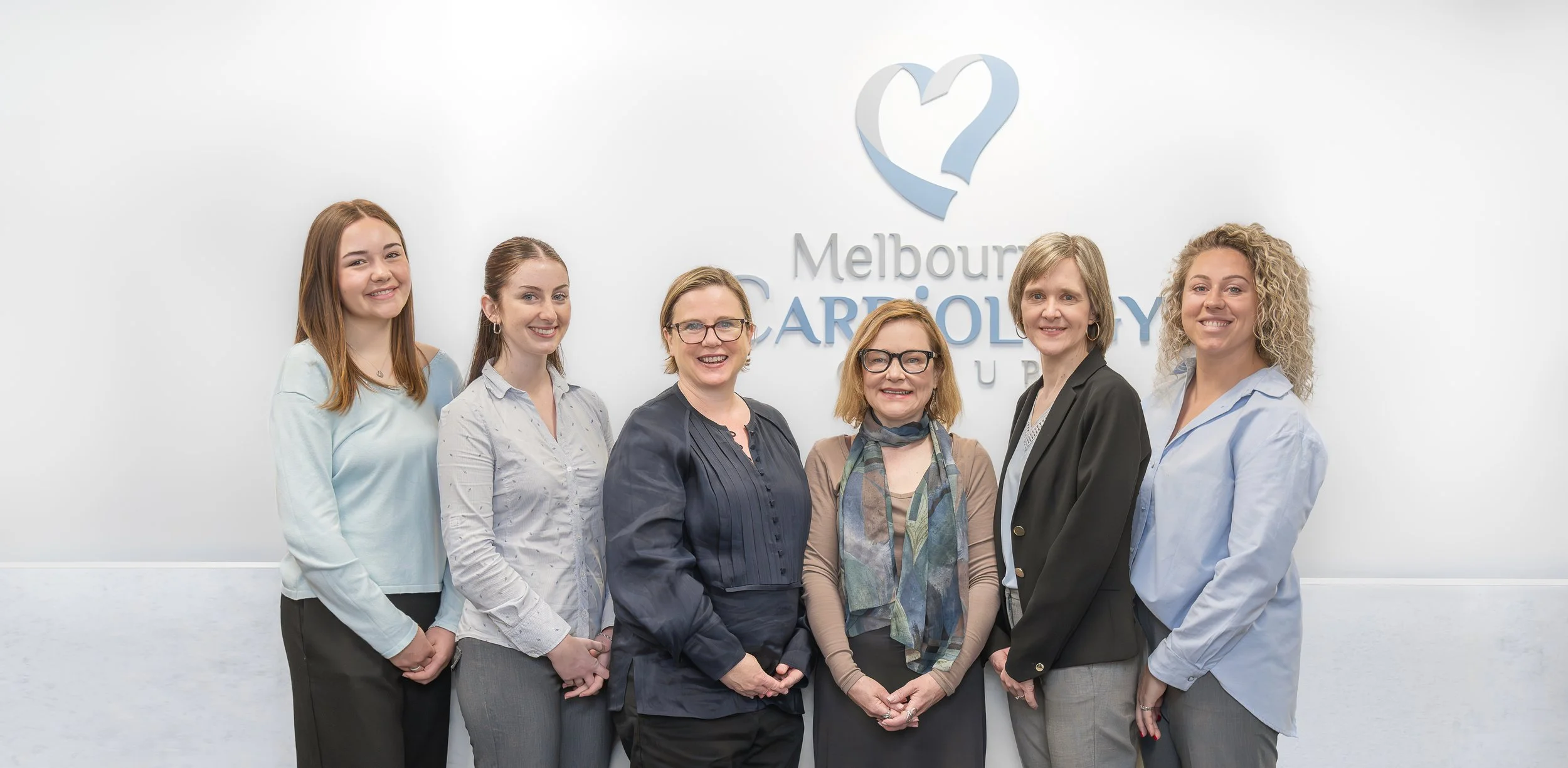 Group of six women standing in front of a white wall with a logo and text that reads 'Melbourne Cardiology'.