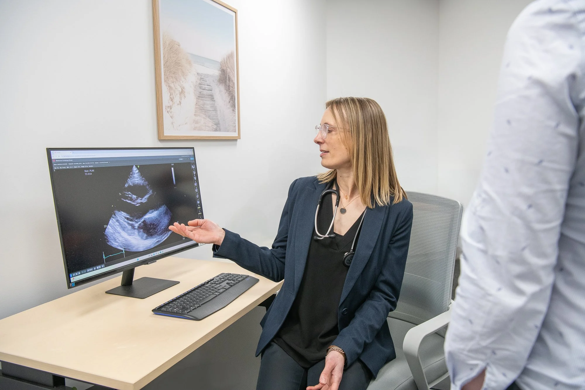 A female doctor in a navy blazer and glasses is explaining an ultrasound image on a computer monitor to a patient. The doctor has a stethoscope around her neck and is seated at a desk with a keyboard and mouse. The patient is standing, partially visible, wearing a white shirt with a pattern. The room has a white wall with a framed picture of a beach scene.