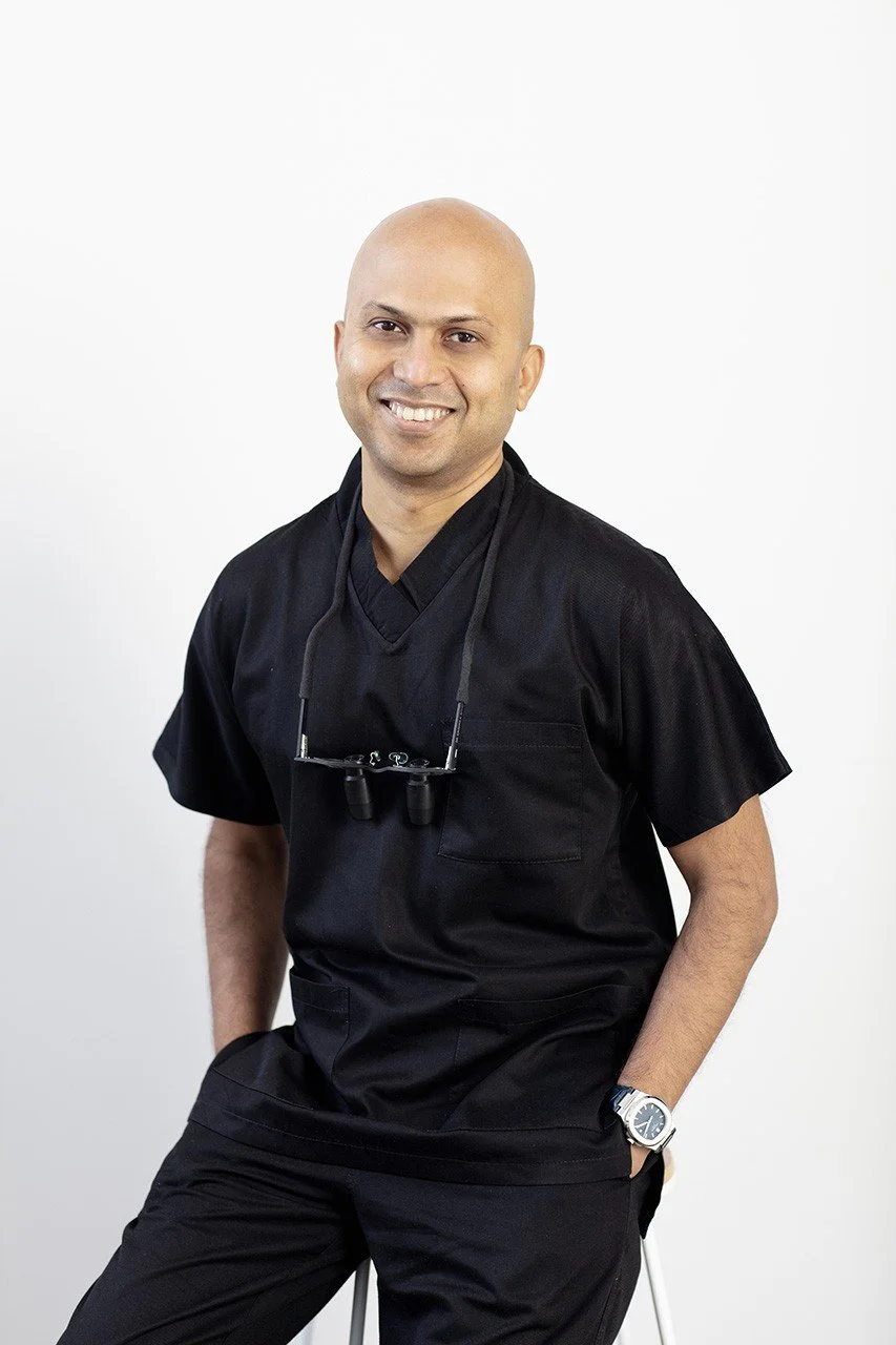 A smiling man in black medical scrubs and a wristwatch, standing against a white background, with a pair of magnifying glasses hanging around his neck.
