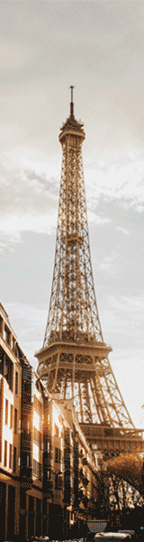 The Eiffel Tower in Paris, France, seen from street level with buildings in the foreground and cloudy sky overhead.