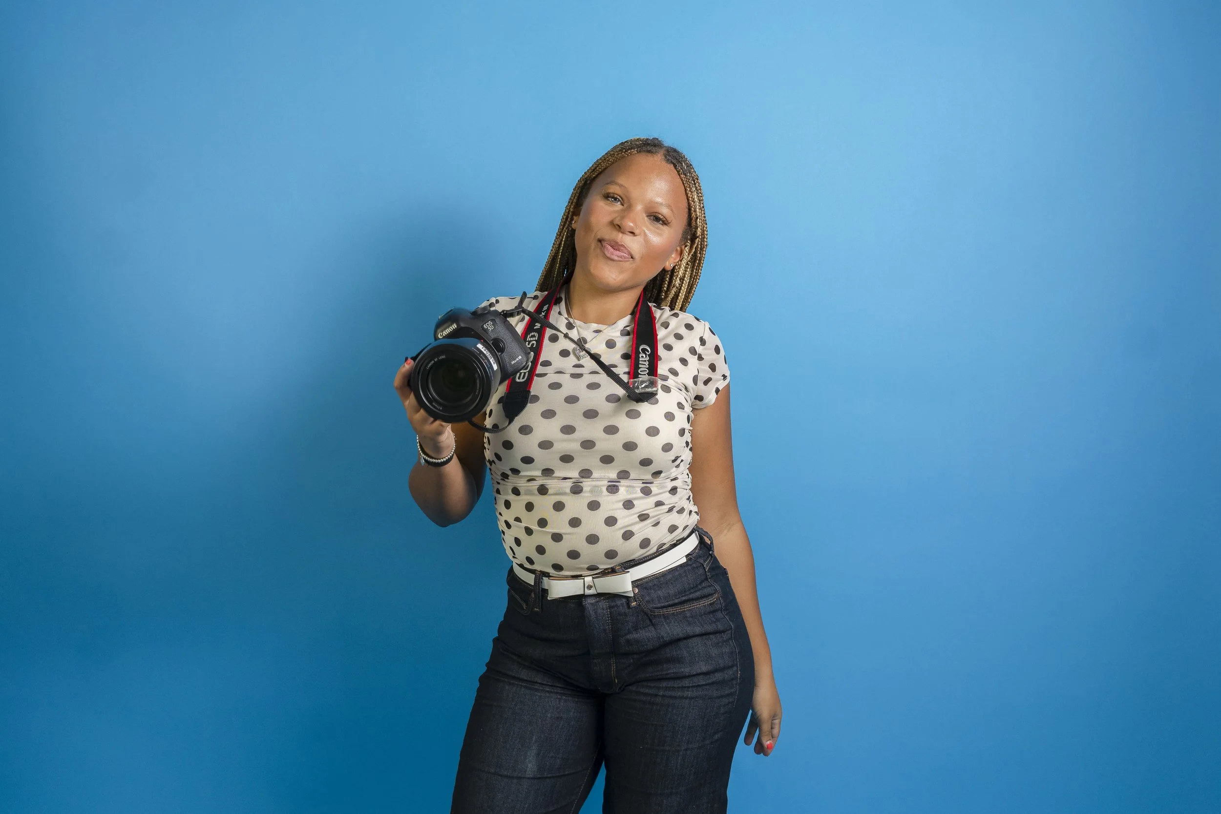 A woman with braided hair holding a Canon camera, standing against a bright blue background.