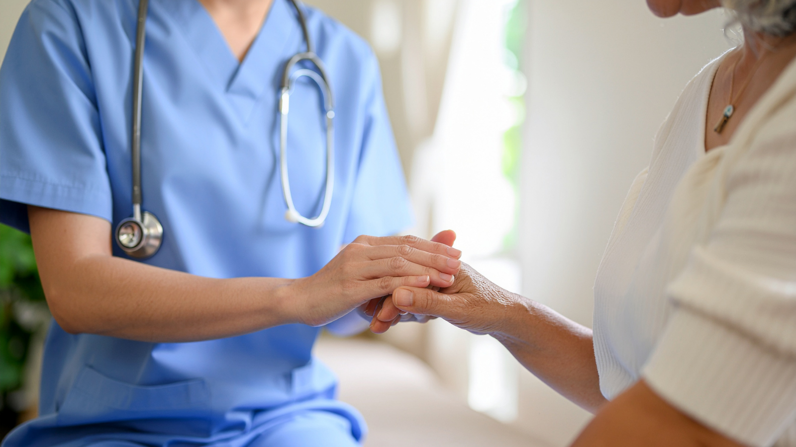 Healthcare worker in blue scrubs and stethoscope holding hands with an elderly woman.