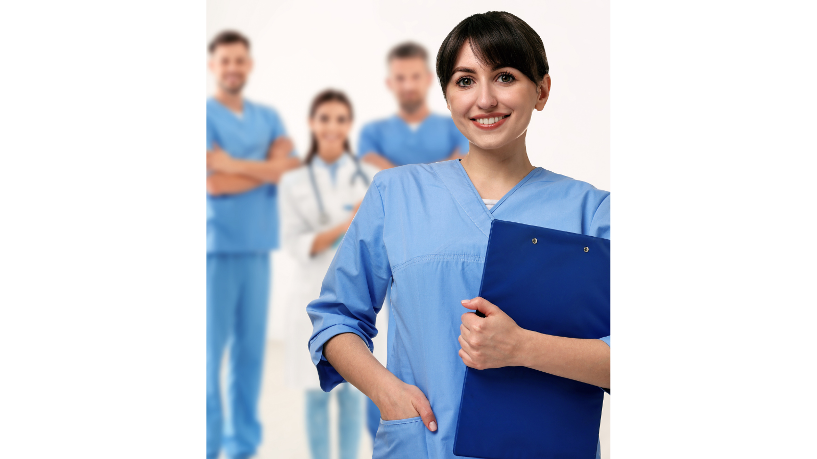 A female healthcare worker with short dark hair, wearing blue scrubs, smiling and holding a blue clipboard, standing in front of three other healthcare professionals in an indoor medical setting.
