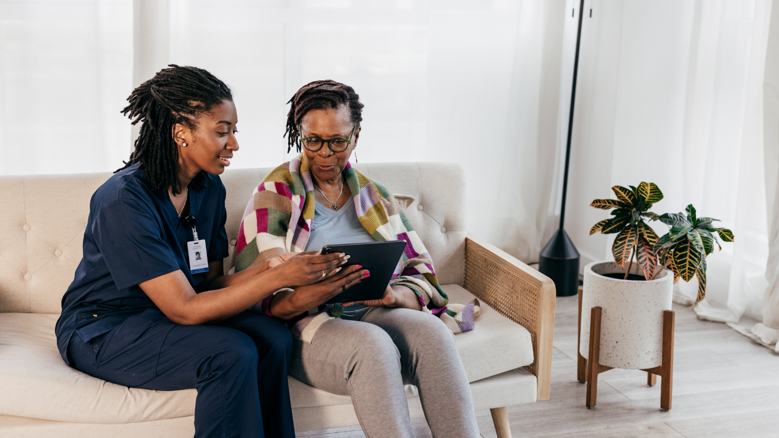 A healthcare professional in navy scrubs showing something on a tablet to a woman sitting on a beige couch in a well-lit living room.