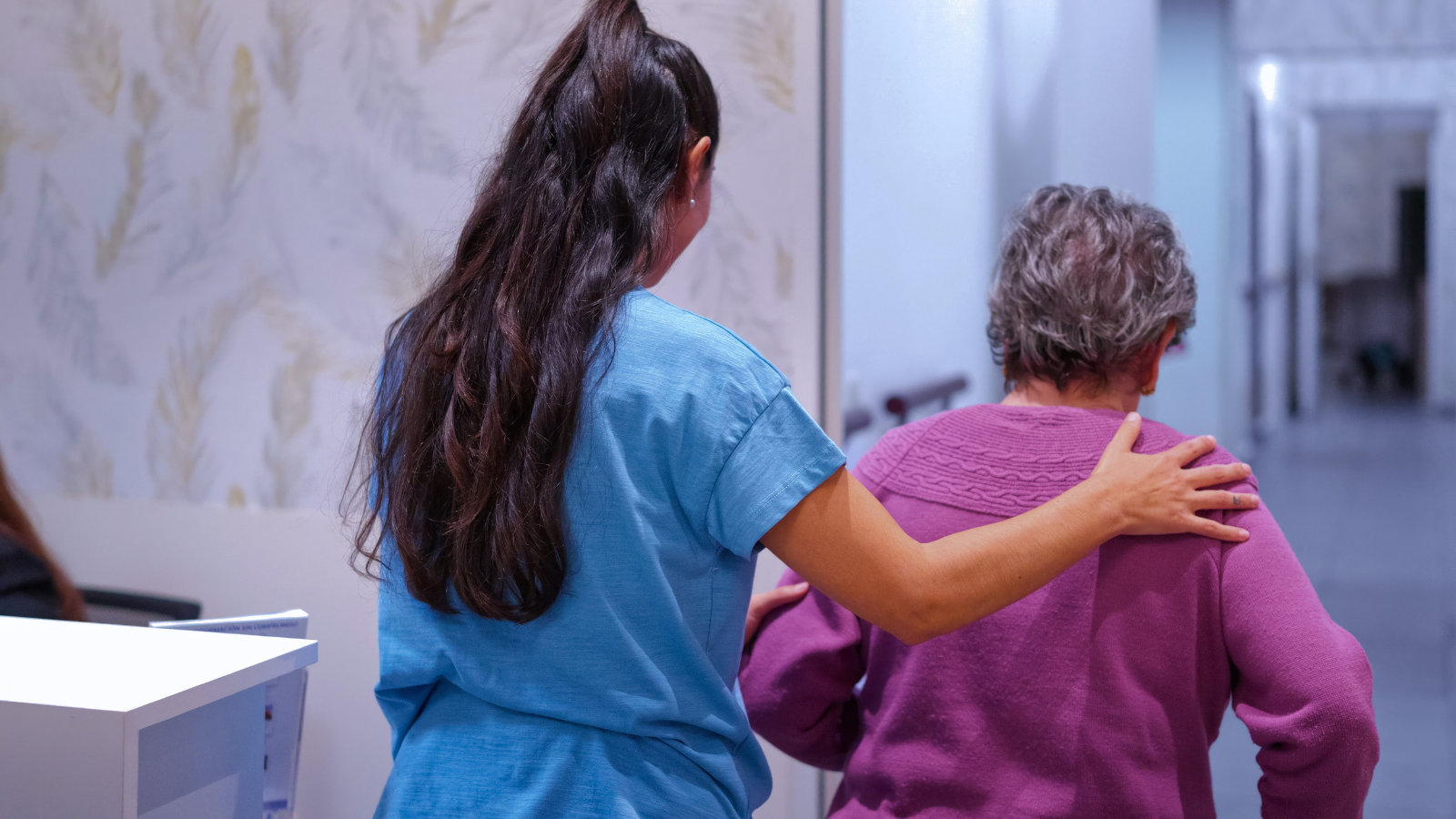 A healthcare worker in blue scrubs gently supports an elderly woman with gray curly hair who is wearing a pink sweater, as they walk down a hospital corridor.
