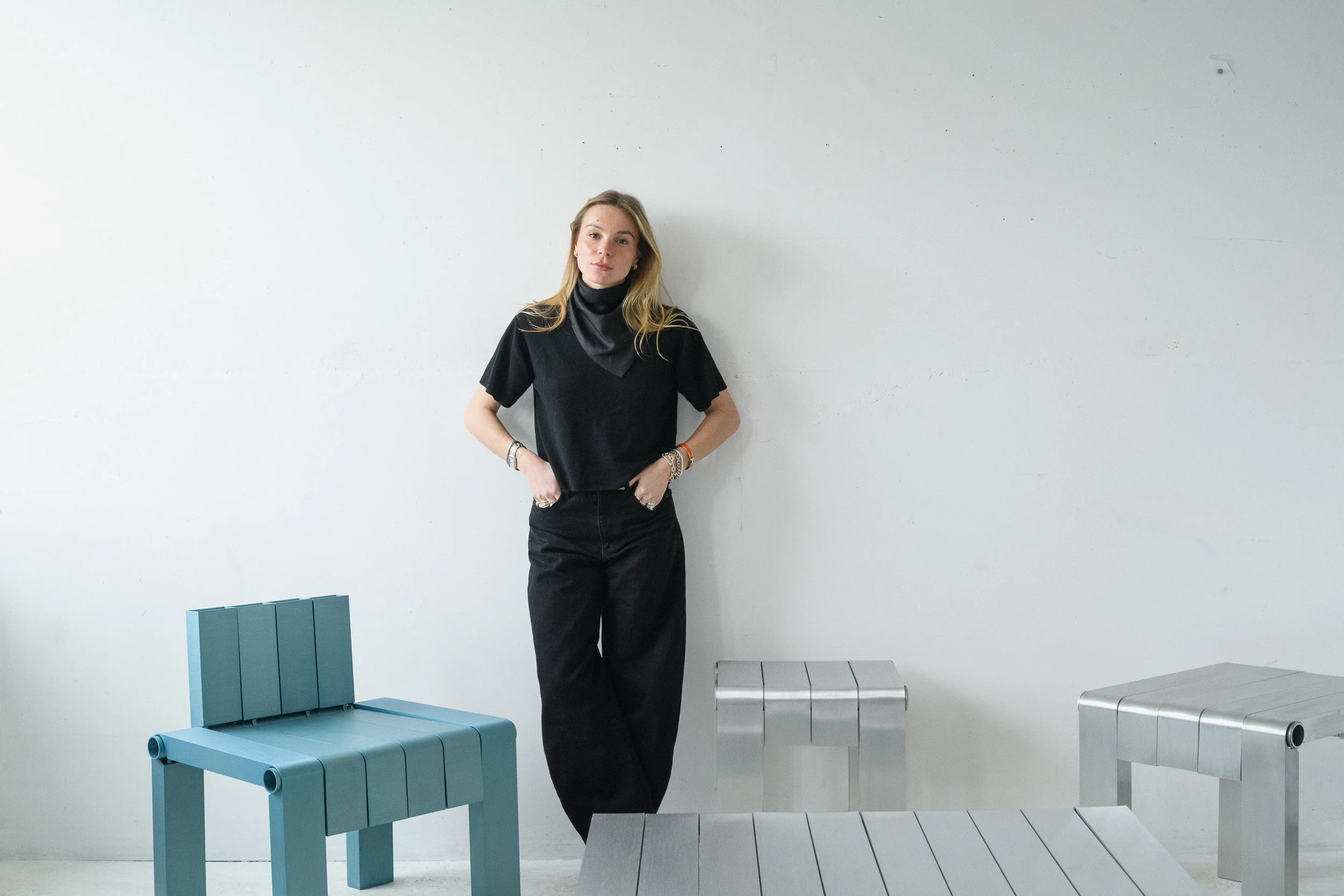 A woman with long blonde hair wearing a black t-shirt, black pants, and multiple bracelets, standing against a plain white wall. There are two modern benches in front, one teal and one gray, with a minimalist design.