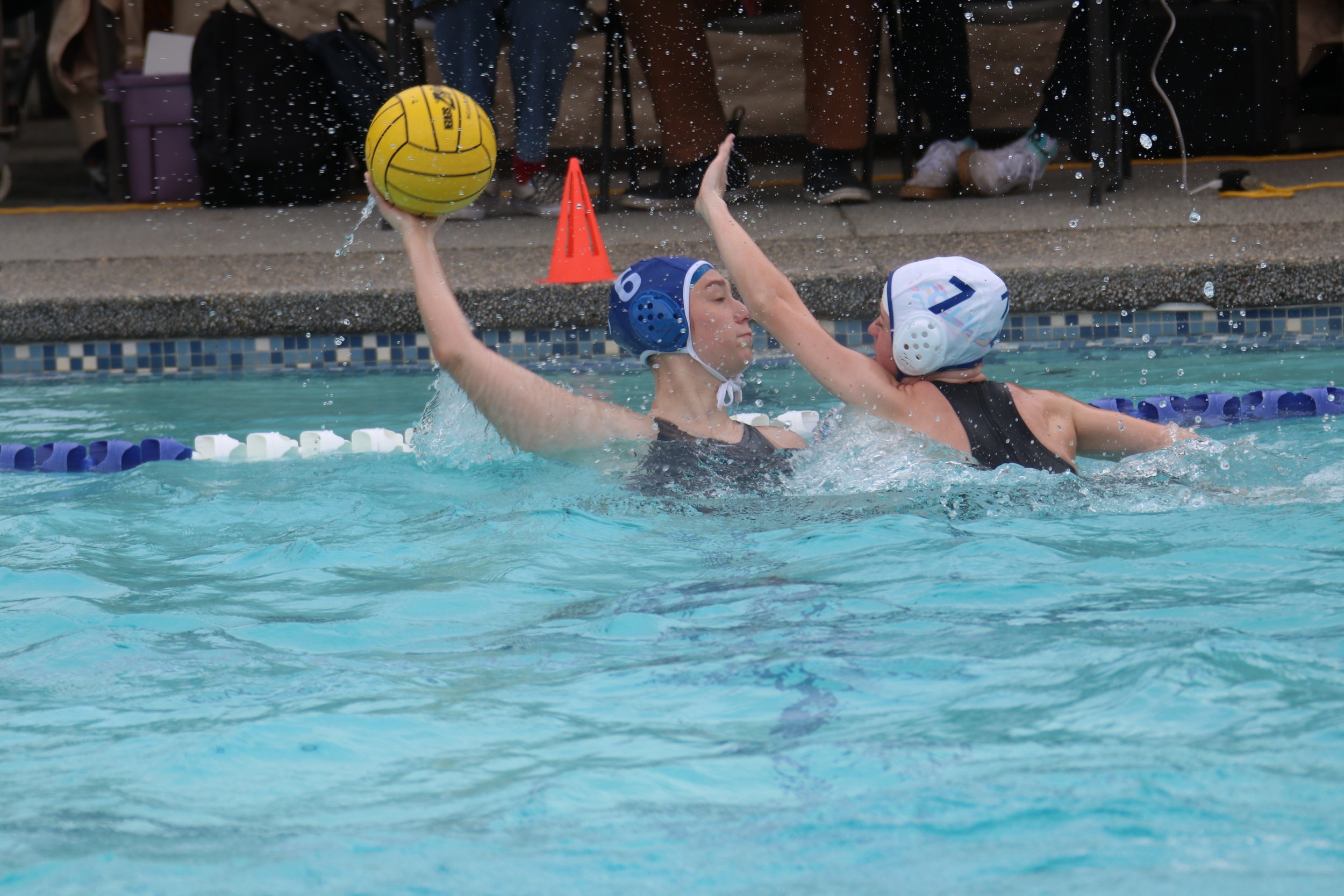 Two water polo players wearing blue caps are competing in a swimming pool. One player, holding a yellow water polo ball, attempts to catch or pass the ball, while the other player is defending. There are spectators sitting behind a barrier in the background.