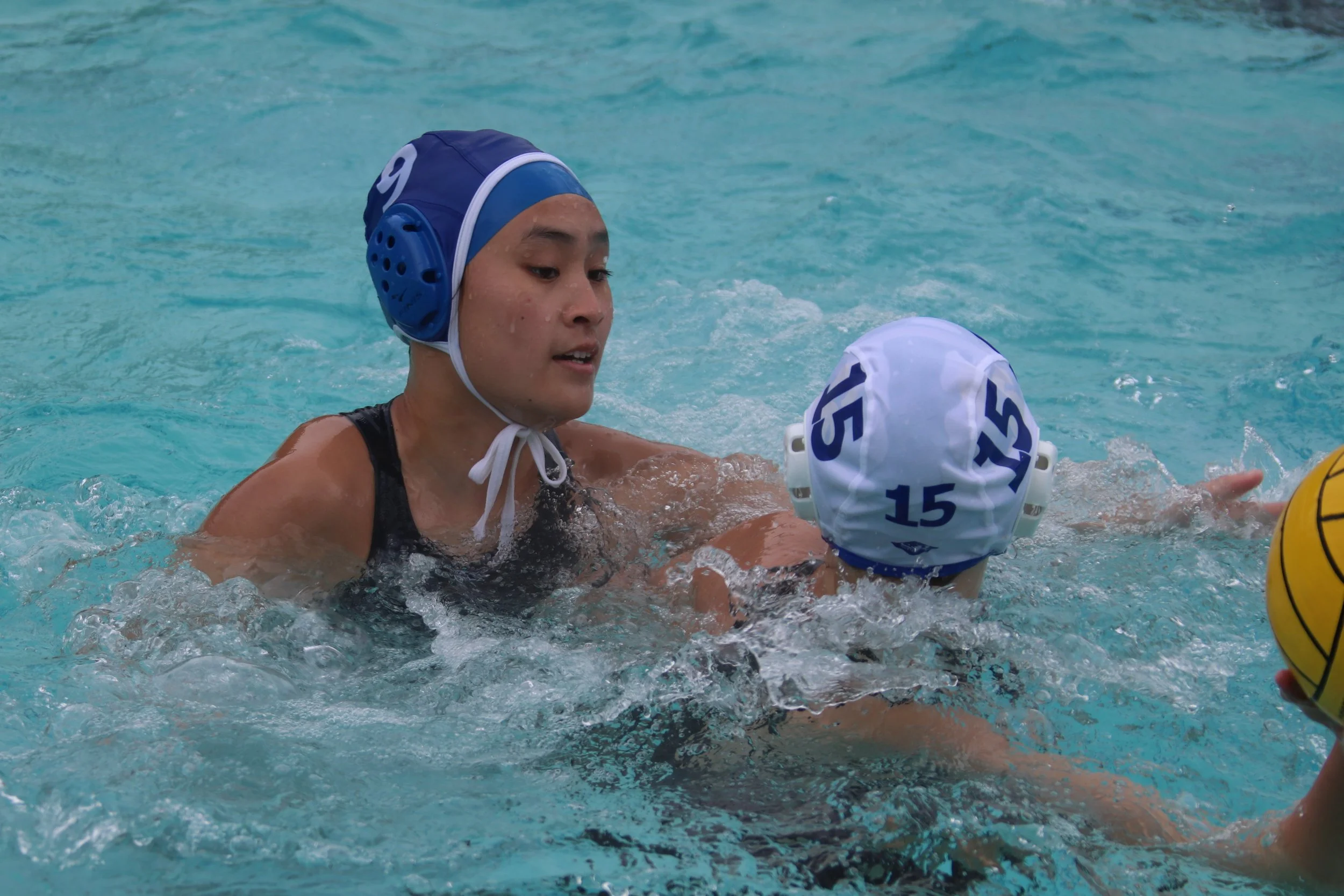 A young water polo player wearing a blue cap with the number 9, in a swimming pool, engaging with another player who has a white cap with the number 15 and the name 'SDS.' The player is in the water, with their face and upper body visible.
