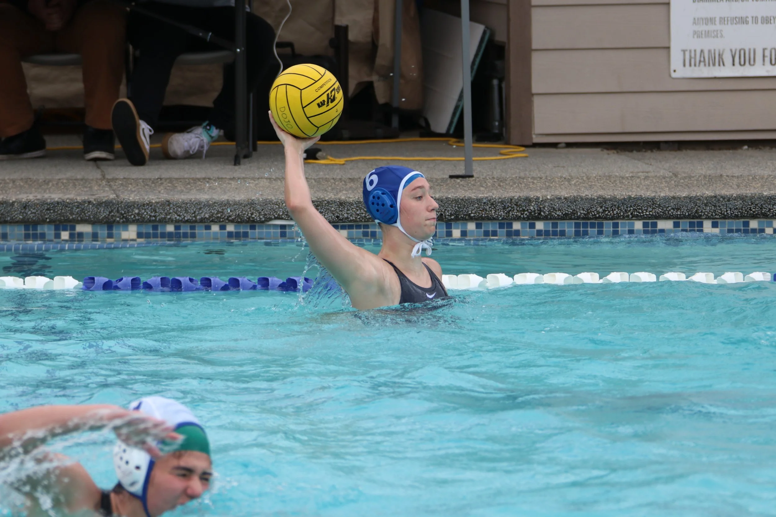 A water polo player in a blue cap and black swimsuit raises a yellow water polo ball above the water, preparing for a pass or shot during a game.