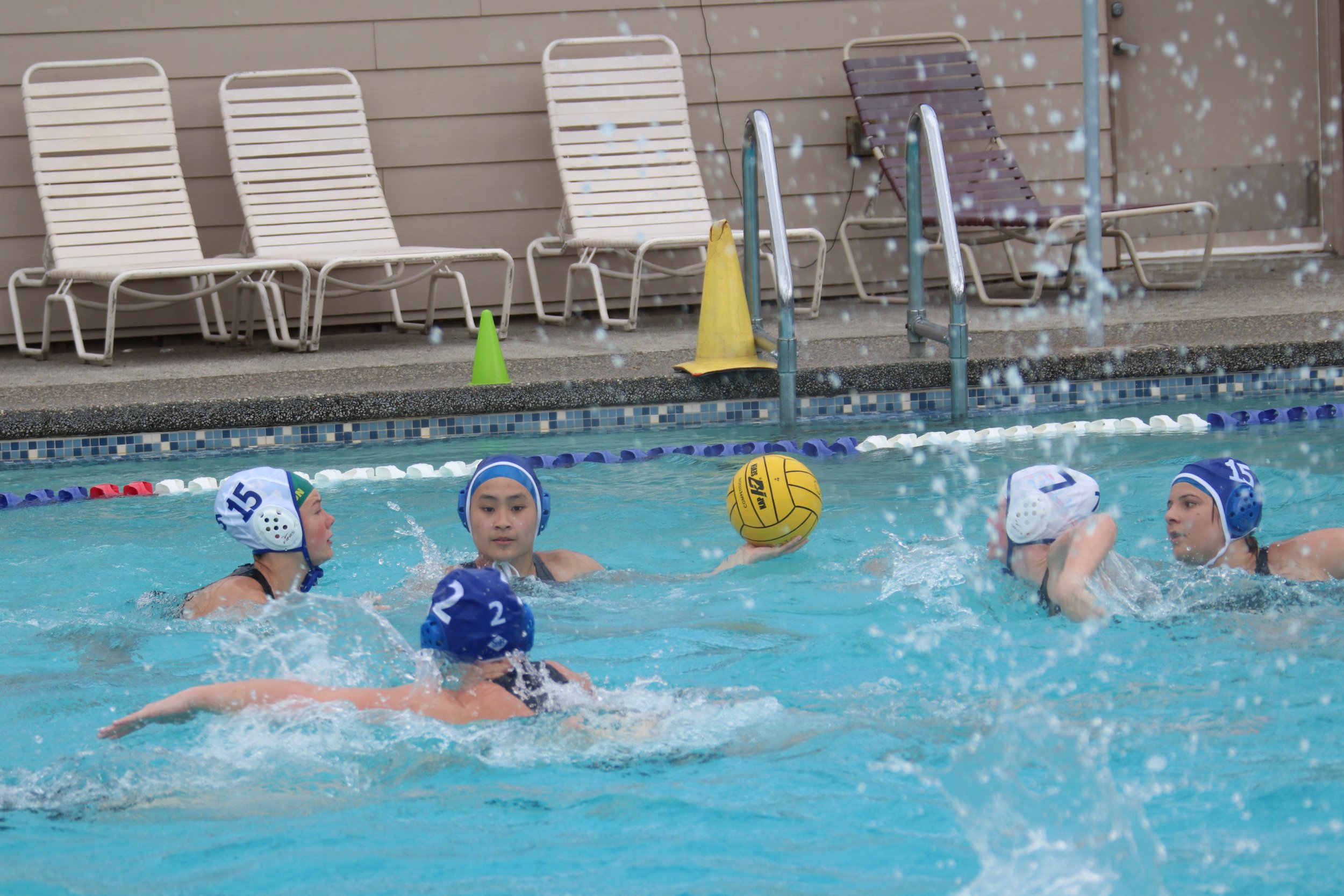 Water polo players in the pool during a game, with chairs, cones, and pool ladder in the background.