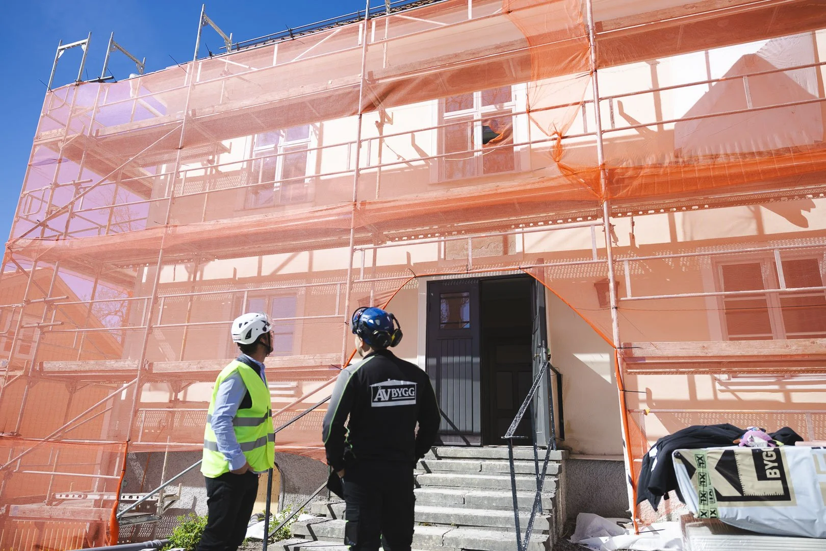 Två medarbetare från ÅV Bygg framför en byggnad inklädd i ställning och väderskydd