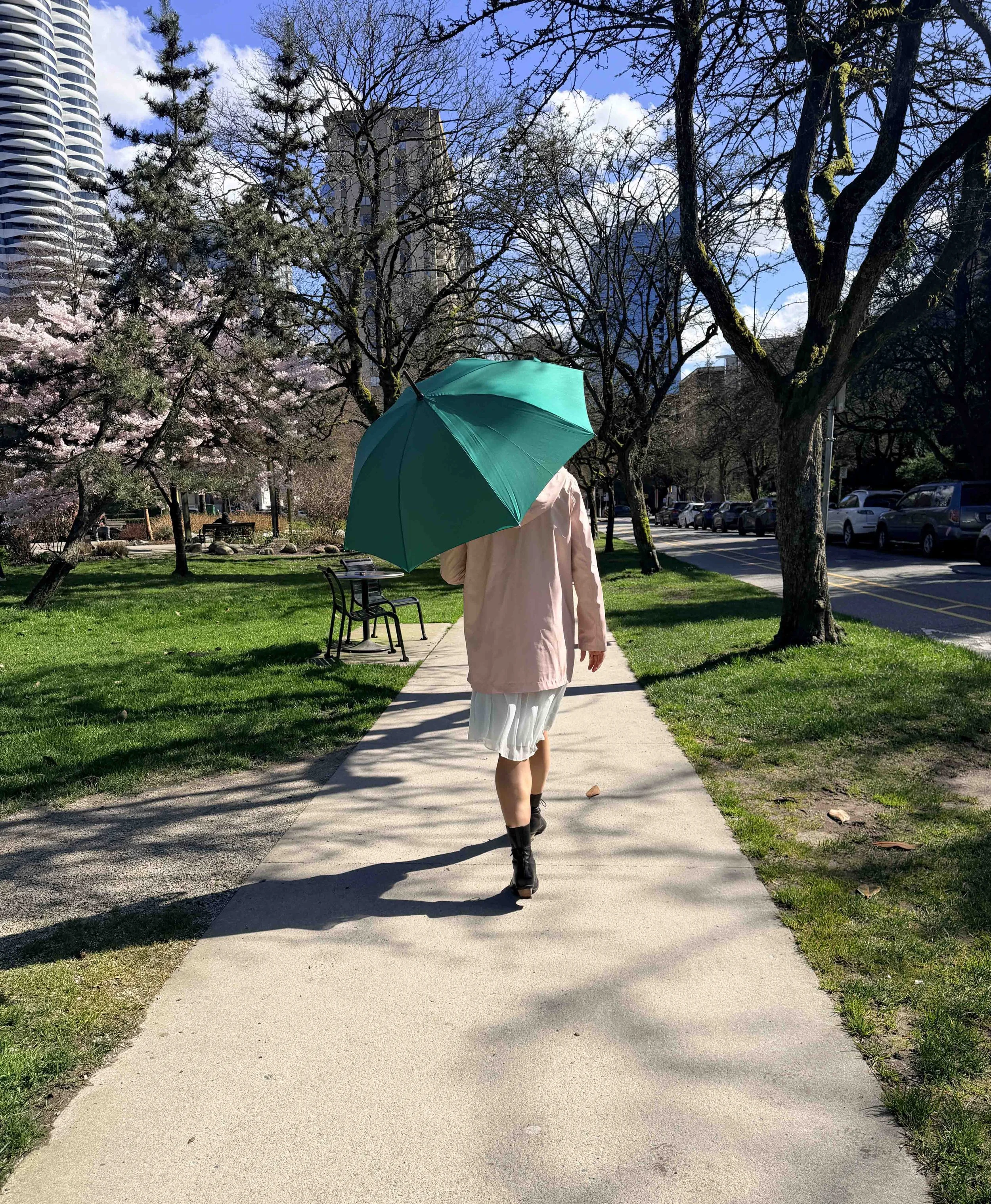 Person walking on a sidewalk holding a green umbrella on a sunny day with trees, a park bench, and buildings in the background.