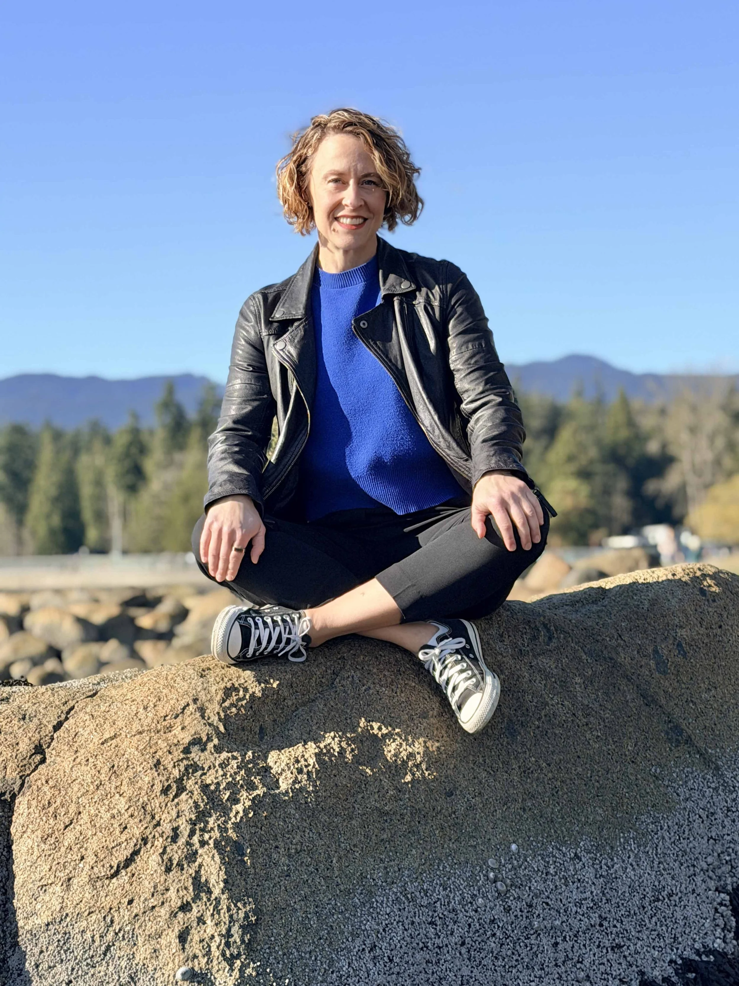 A woman with short curly blonde hair sitting cross-legged on a large rock outdoors, smiling, with mountains and trees in the background under a clear blue sky.