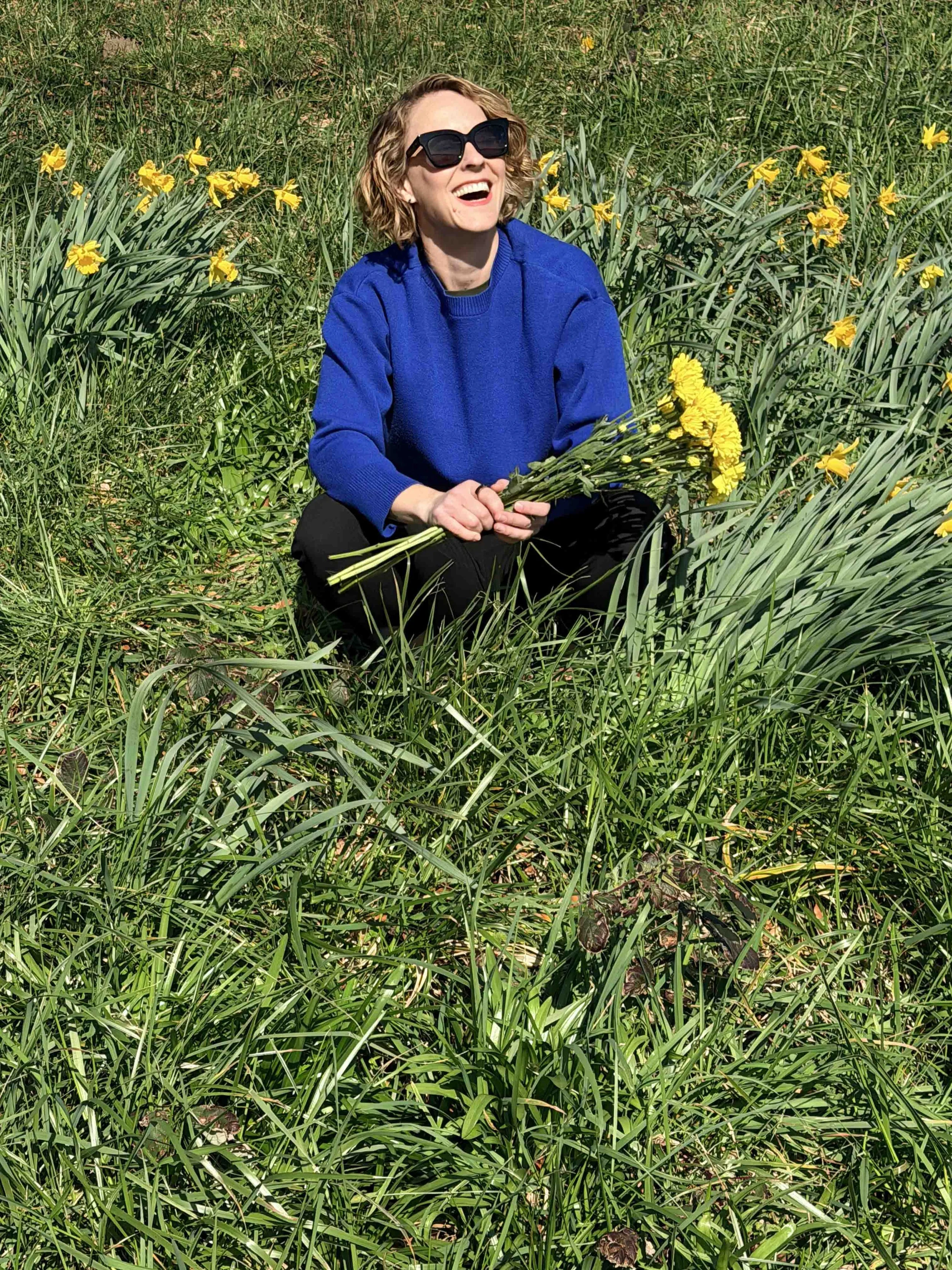 Woman sitting in a field of yellow flowers, holding a bouquet, wearing a blue sweater and sunglasses, smiling and enjoying the sunny day.