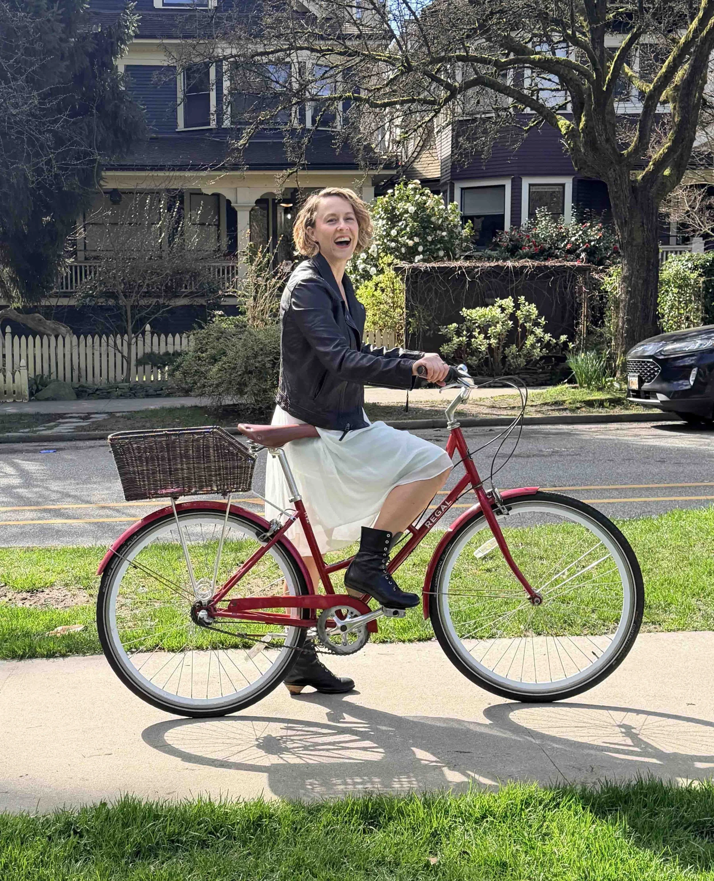 A woman riding a red bicycle on a sidewalk in a residential neighborhood. She is smiling, wearing a black leather jacket, a white dress, and black boots. The background features trees and houses.