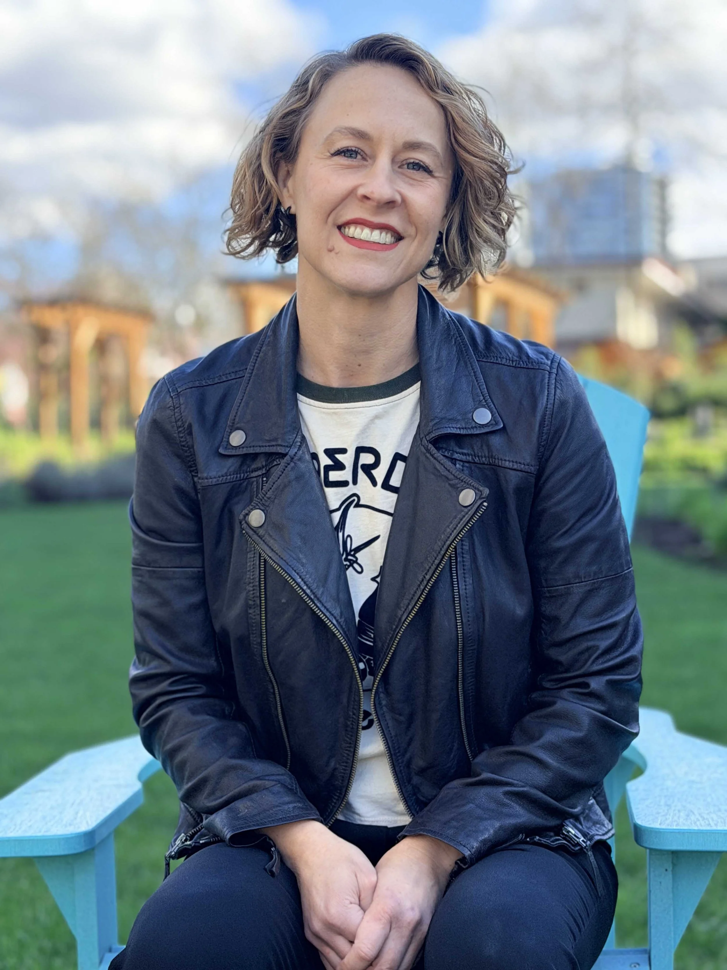 A woman with short curly hair sitting outdoors on a blue chair, smiling at the camera, wearing a black leather jacket and a graphic t-shirt, with a garden and buildings in the background.