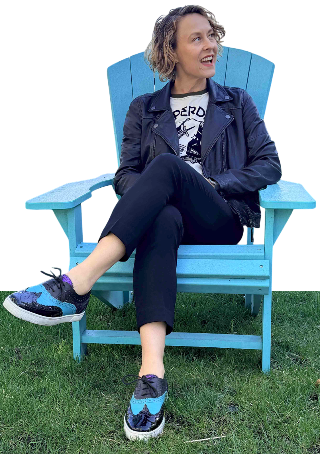 A woman with curly blonde hair sitting on a bright blue Adirondack chair outside on grass, wearing a black leather jacket, a white t-shirt with a graphic, black pants, and colorful black and blue sneakers. She is smiling and looking to her right.