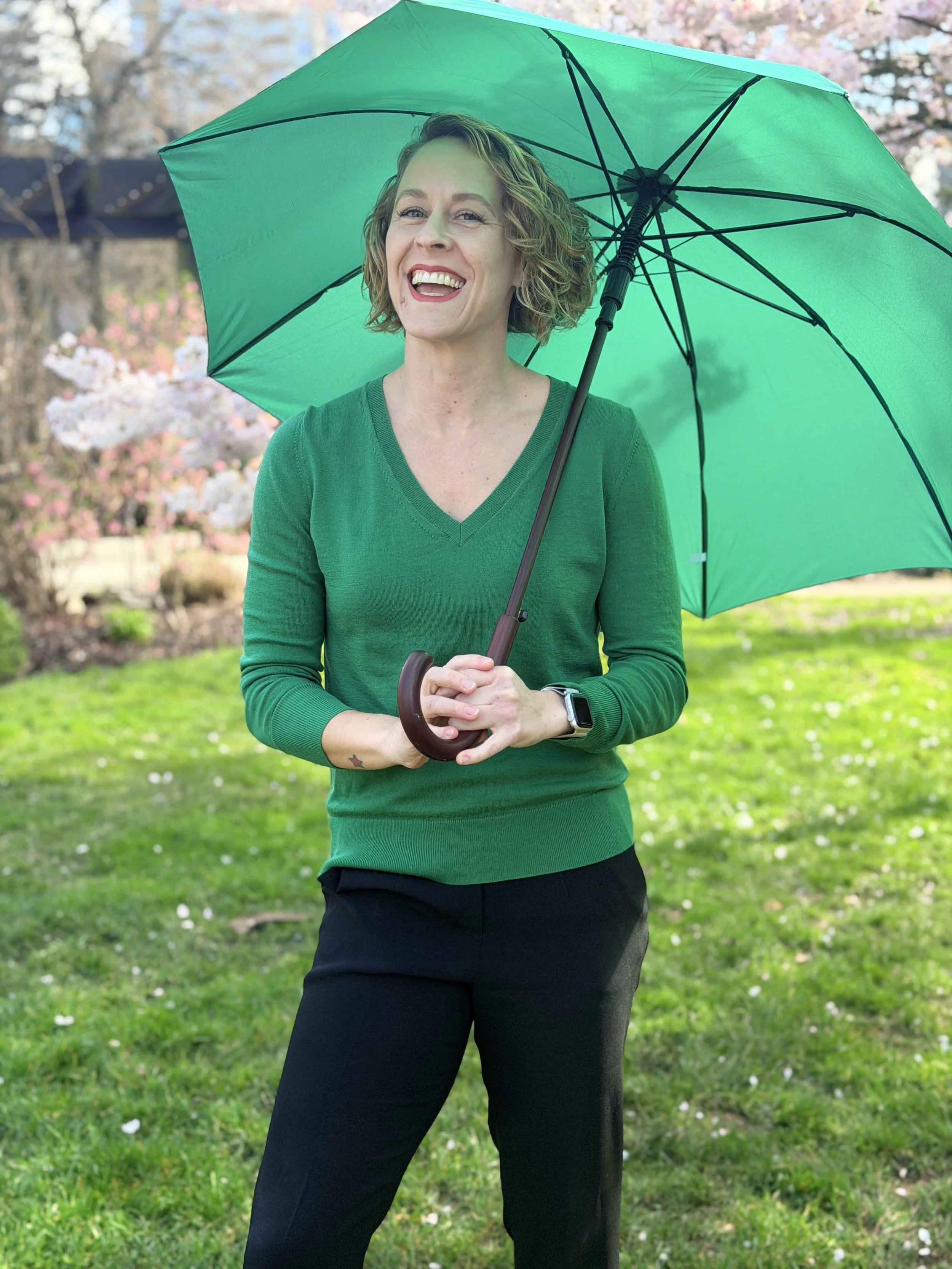 A smiling woman holding a green umbrella outdoors in a park with blooming trees and green grass.