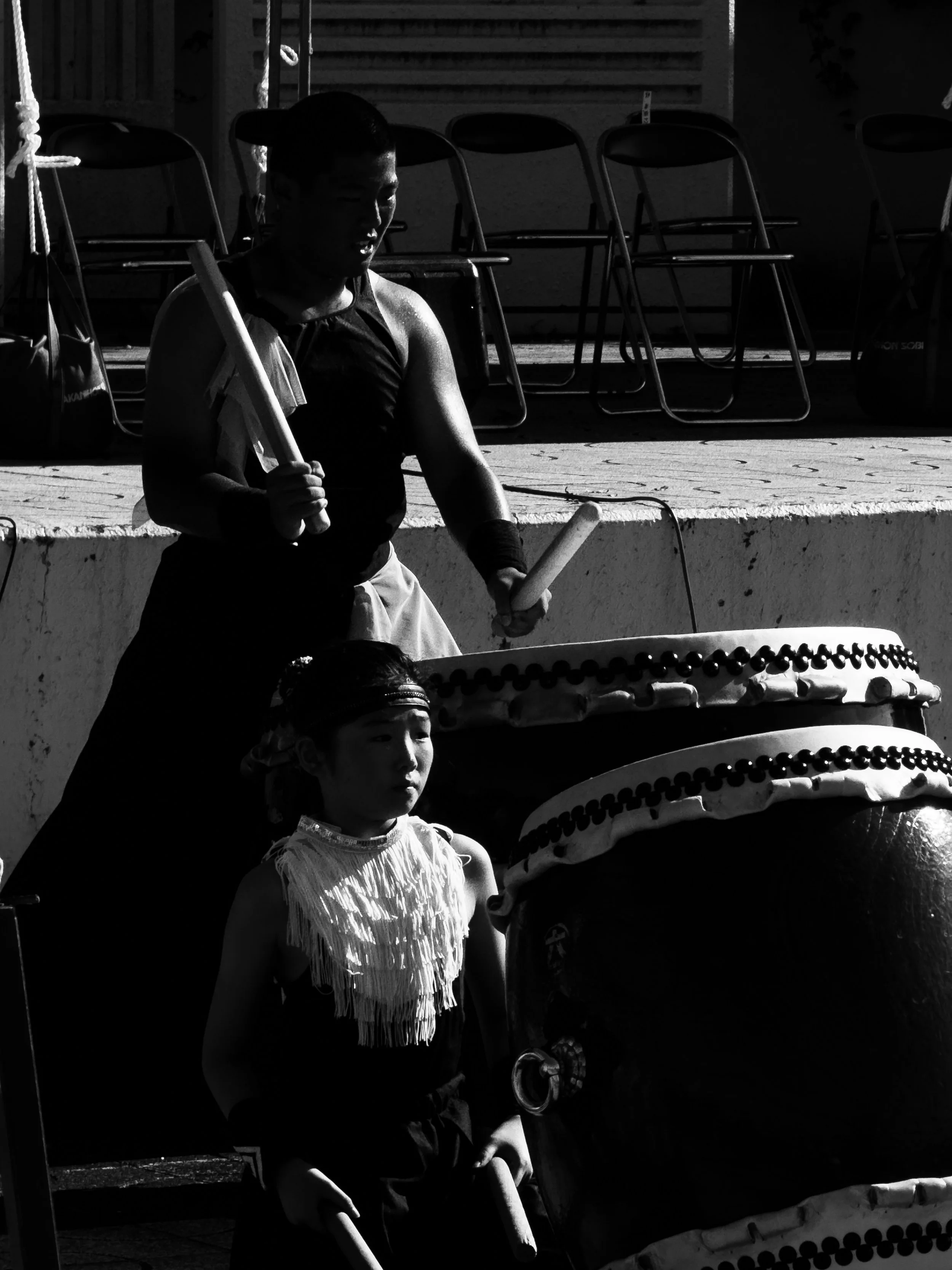Taiko performer holding drumsticks beside large drums in black and white.