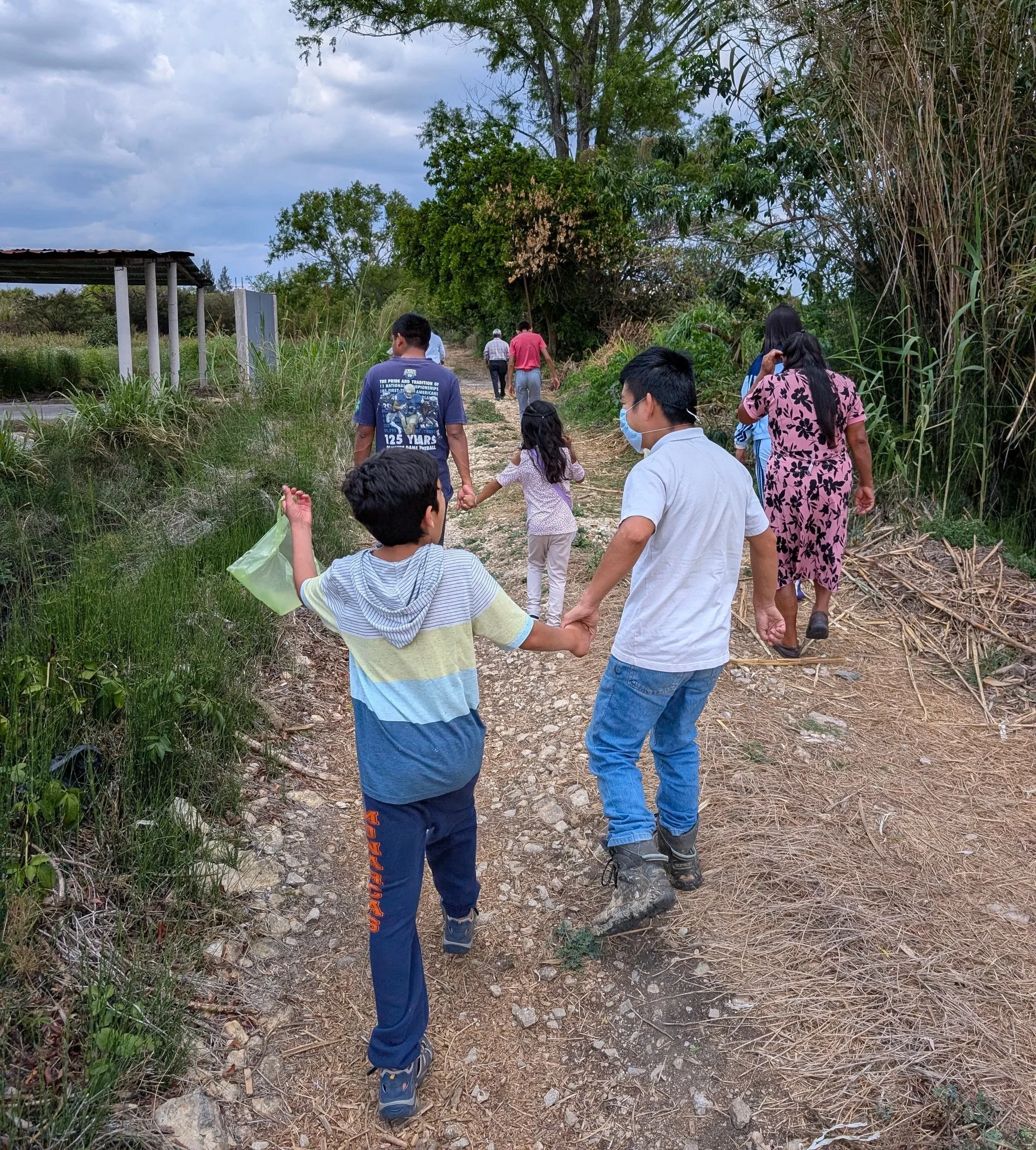 People walking hand in hand on a dirt trail through a natural outdoor setting with trees and bushes, under a cloudy sky.