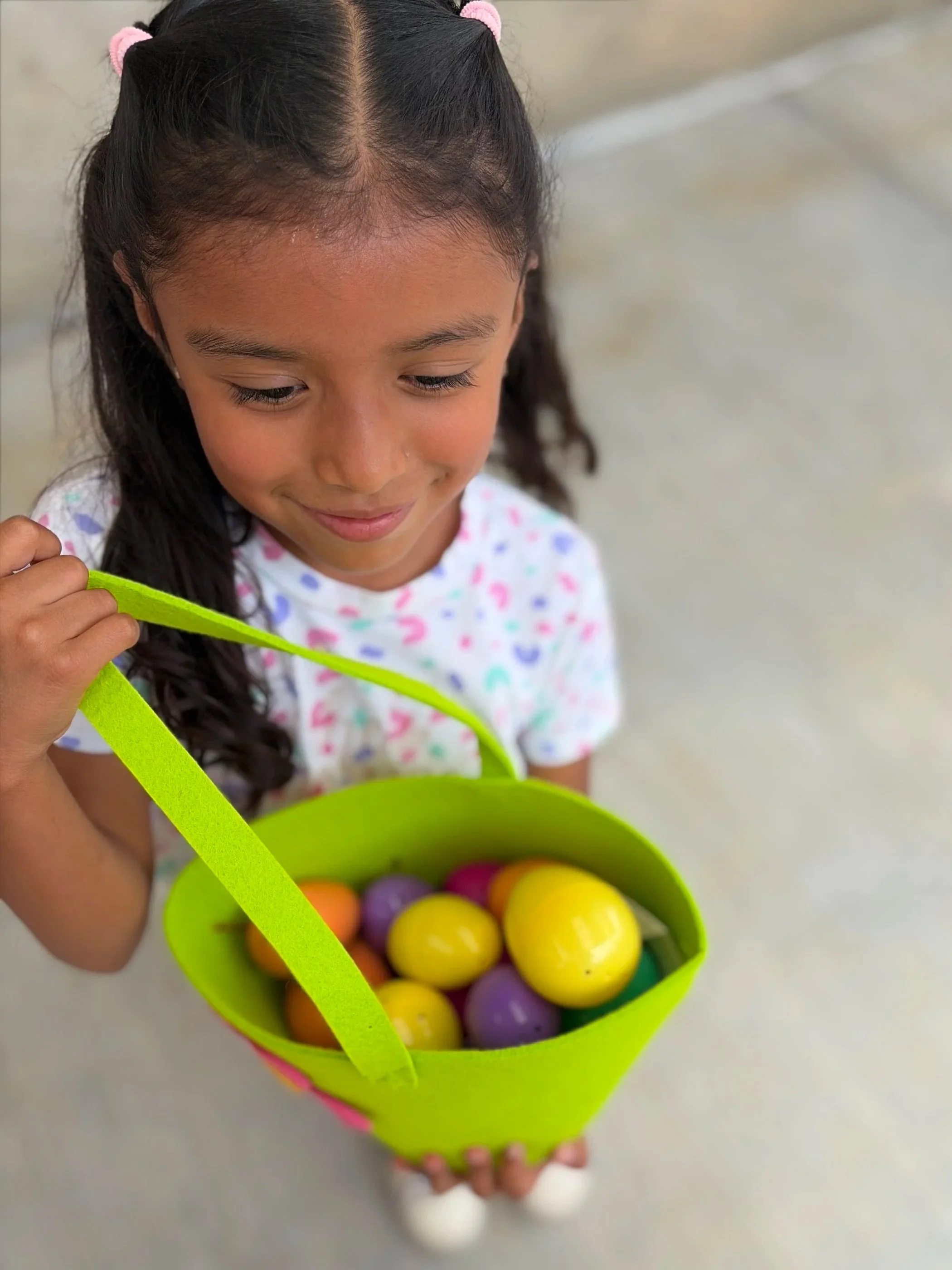 A young girl with dark hair in pigtails wearing pink hair ties, holding a green basket filled with colorful plastic Easter eggs.
