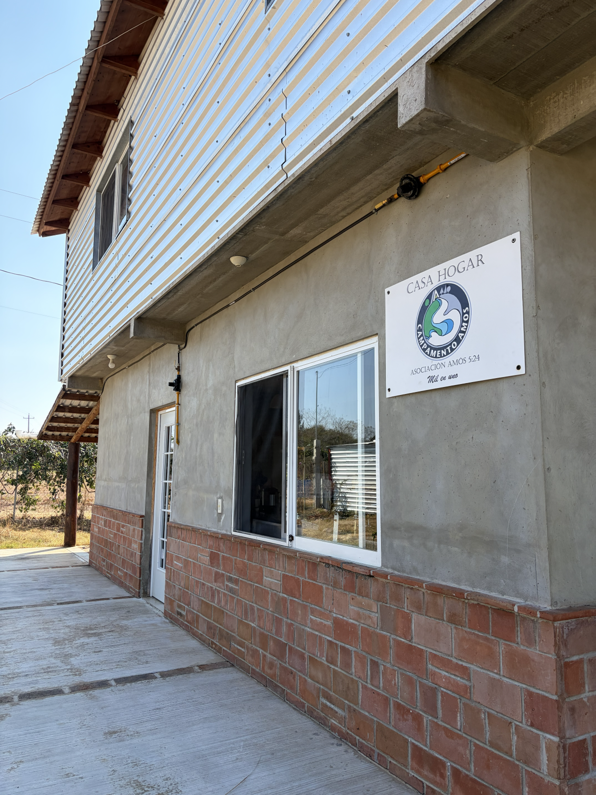 Front view of a house with a small porch, a window, a door, and a large sign that reads 'Casa Hogar'. The house has a concrete and brick lower section, a gray concrete upper section, and a metal roof. There are electrical wires and a meter on the wall.