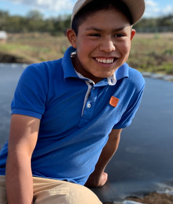 A boy smiling outdoors near a body of water, wearing a beige cap and a blue polo shirt.