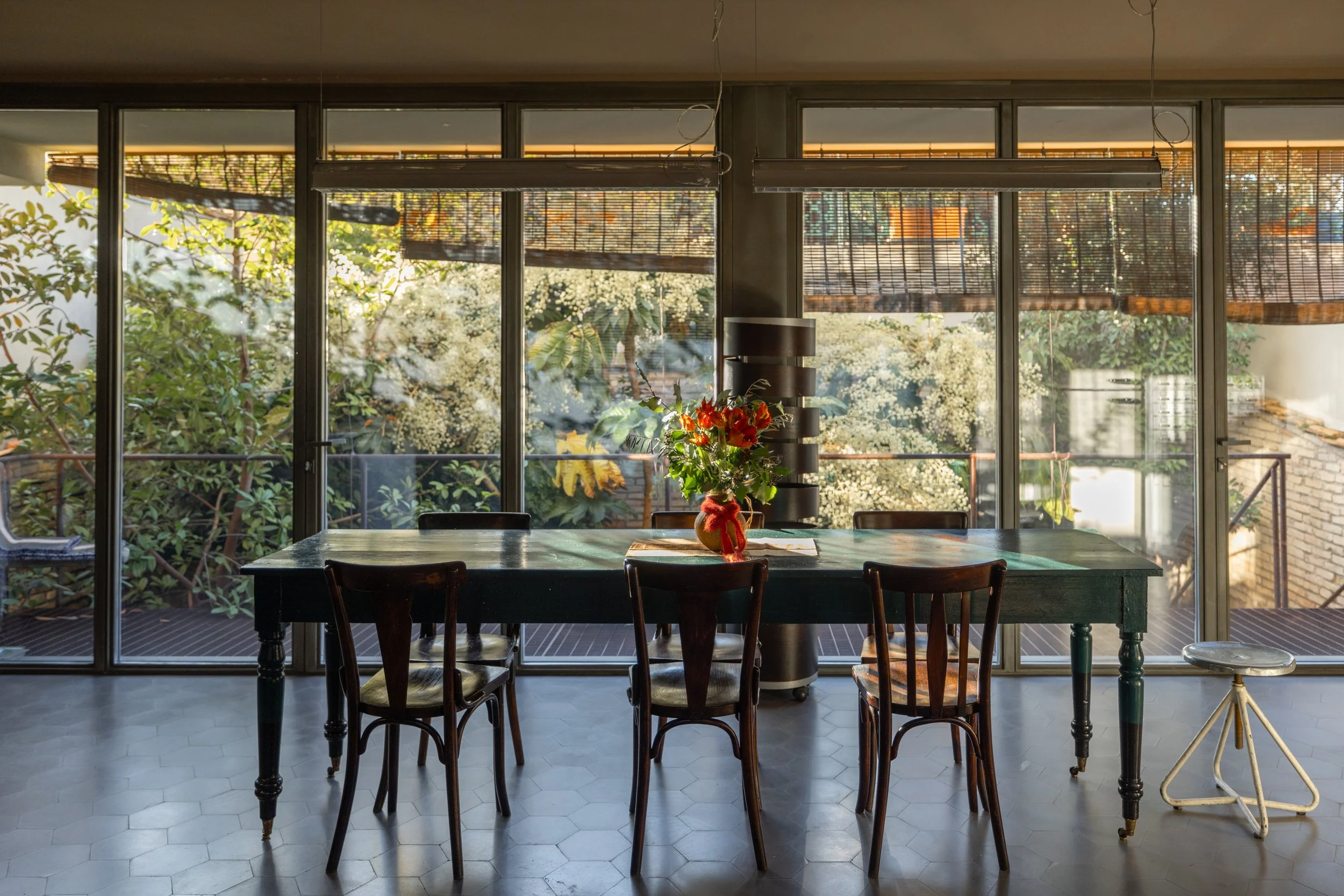 Dining area featuring a large wooden table with chairs, a flower vase centerpiece, and a background of glass sliding doors with bamboo blinds opening to a lush outdoor garden.