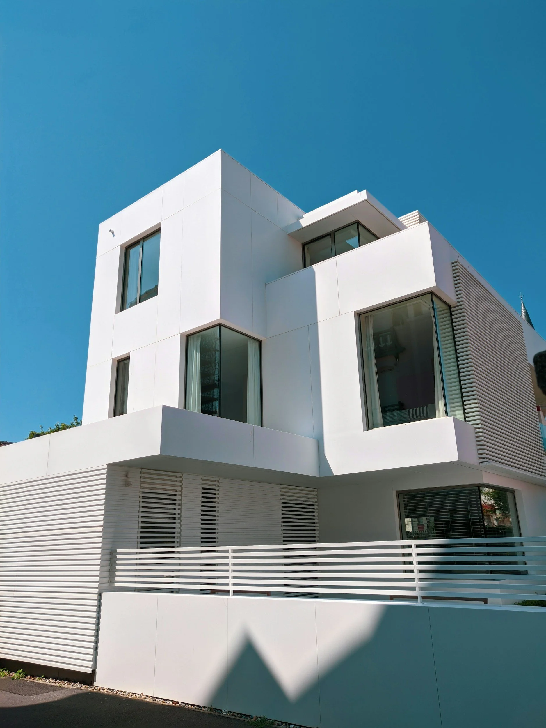 Modern white building with large glass windows and horizontal slatted panels, under a clear blue sky.