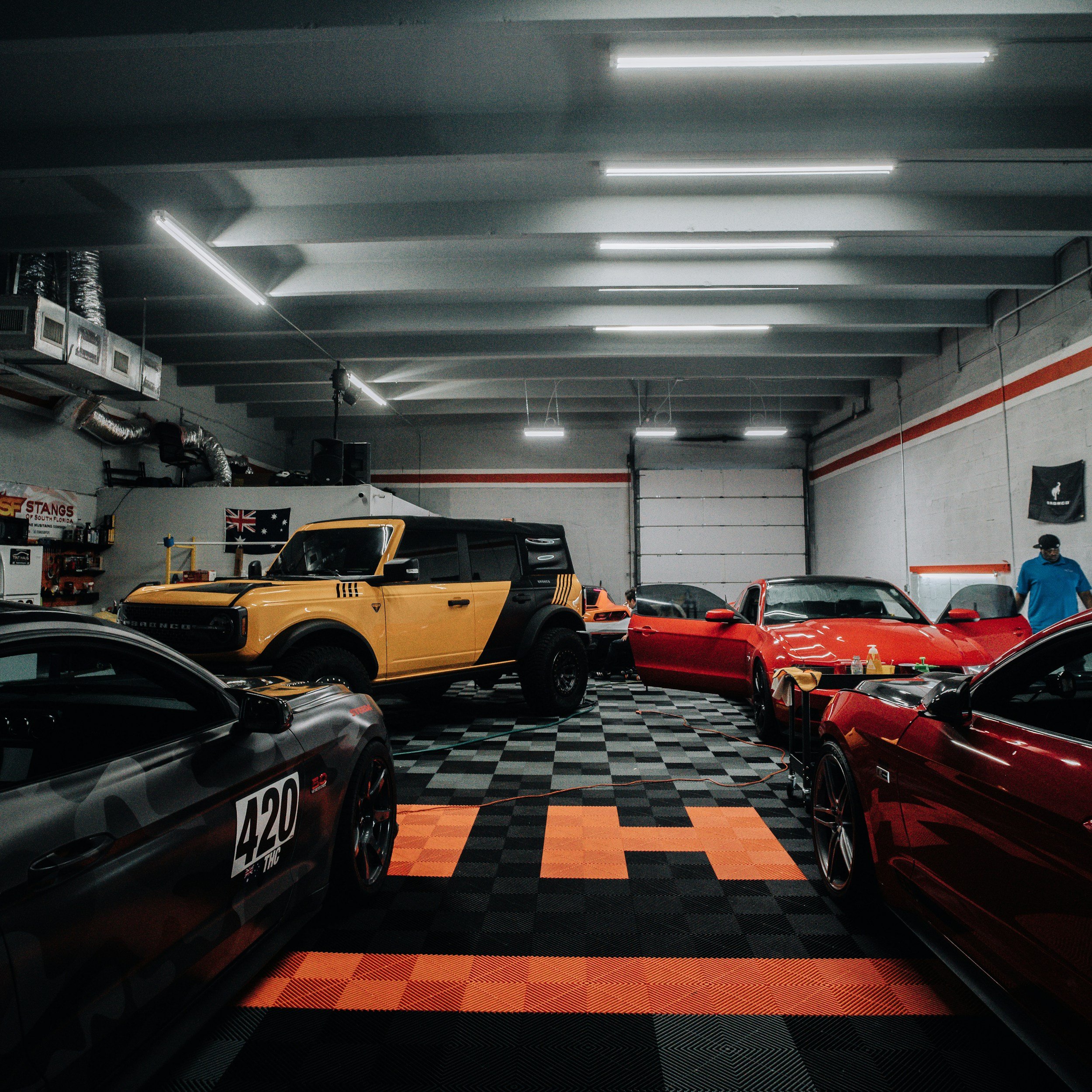 Indoor garage with racing cars, checkered floor, exposed ducts, fluorescent ceiling lights, and a man in a blue shirt.