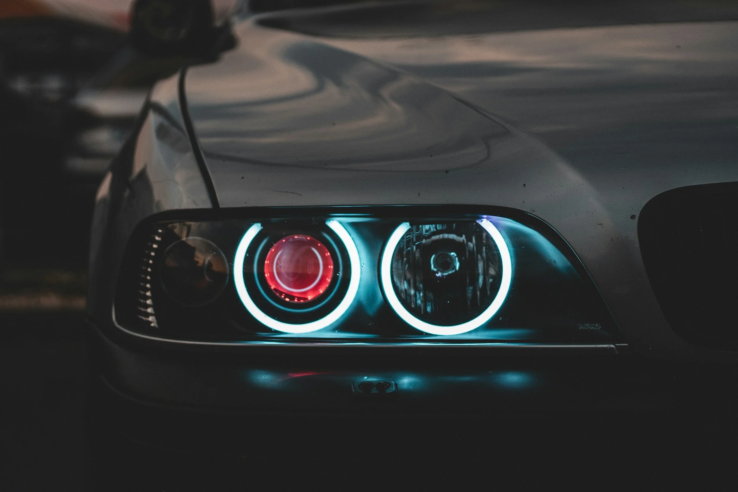 Close-up of a car's front headlight with blue and red illuminated rings in a dark environment.