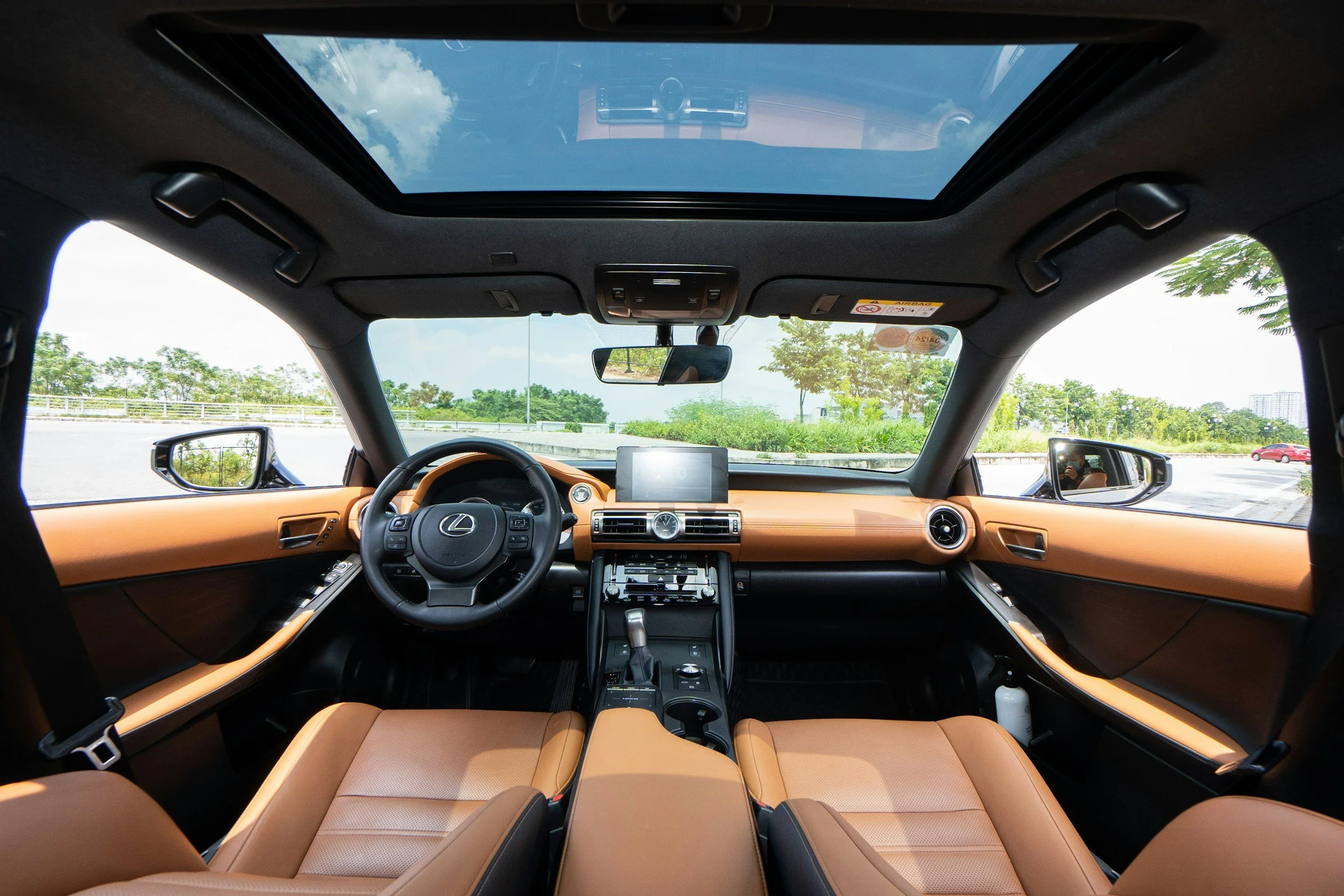 Interior of a luxury car with a panoramic sunroof, tan leather seats, a dashboard with a screen, and a view of blue sky and green trees outside.