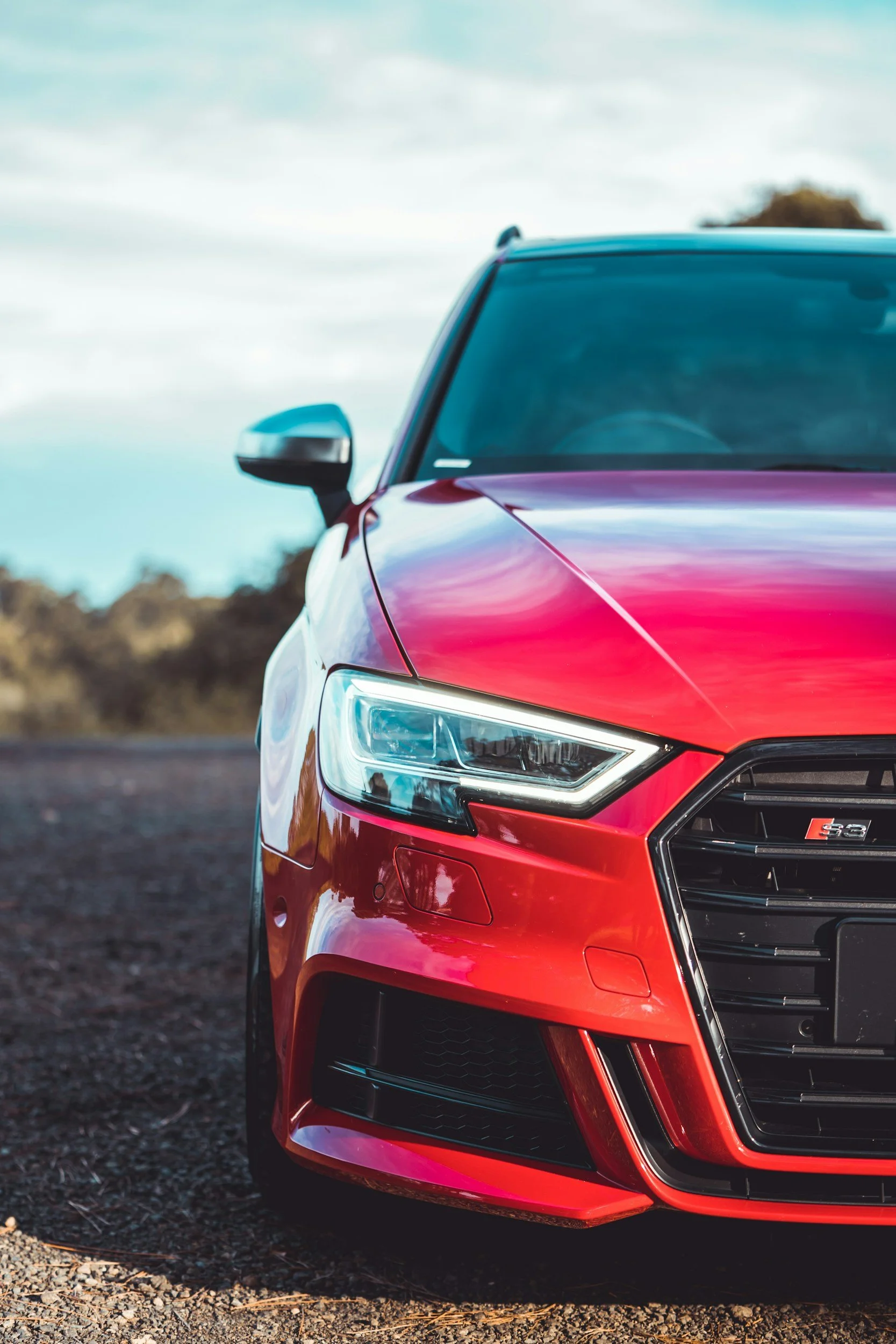 Close-up of the front of a red Audi S3 car with black grille and headlight, parked on gravel outdoor.