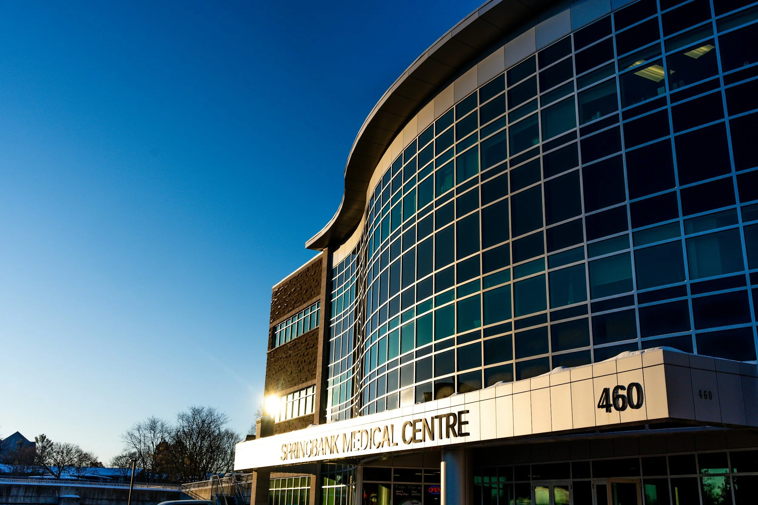 Modern curved glass building with white signage reading "Springbank Medical Centre" and the street number 460, sunlight reflecting off the windows, trees without leaves in the background, clear blue sky.