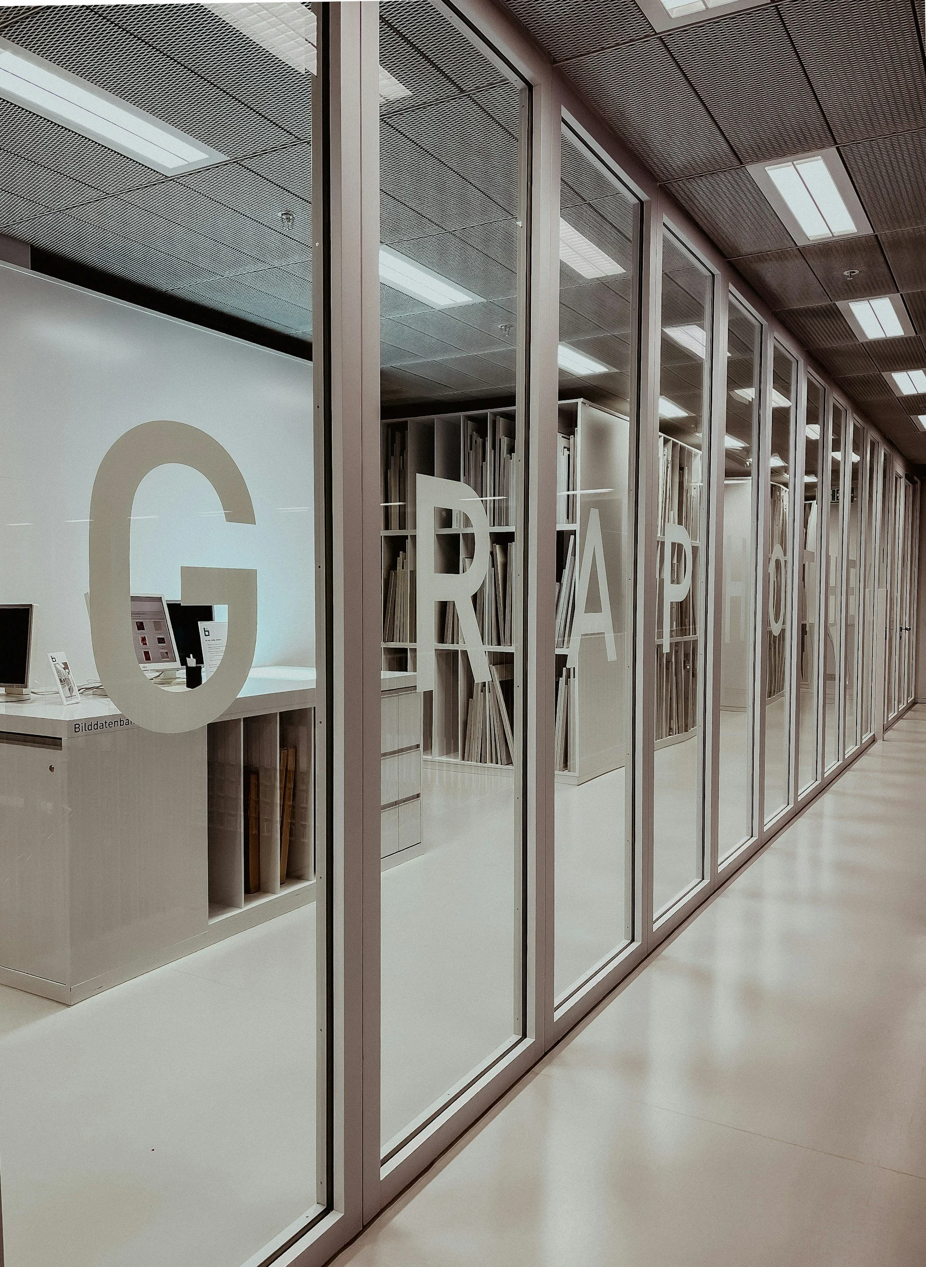 A modern office space with glass walls, containing shelves filled with books and documents, and a white desk with electronic devices, illuminated by ceiling lights.
