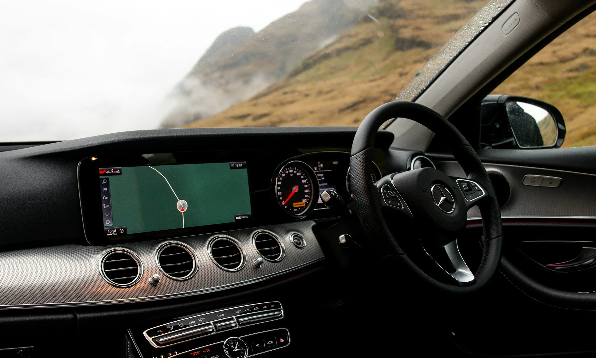 Inside view of a Mercedes-Benz car dashboard with a GPS navigation screen, speedometer, and air vents, while driving through a foggy mountain landscape.