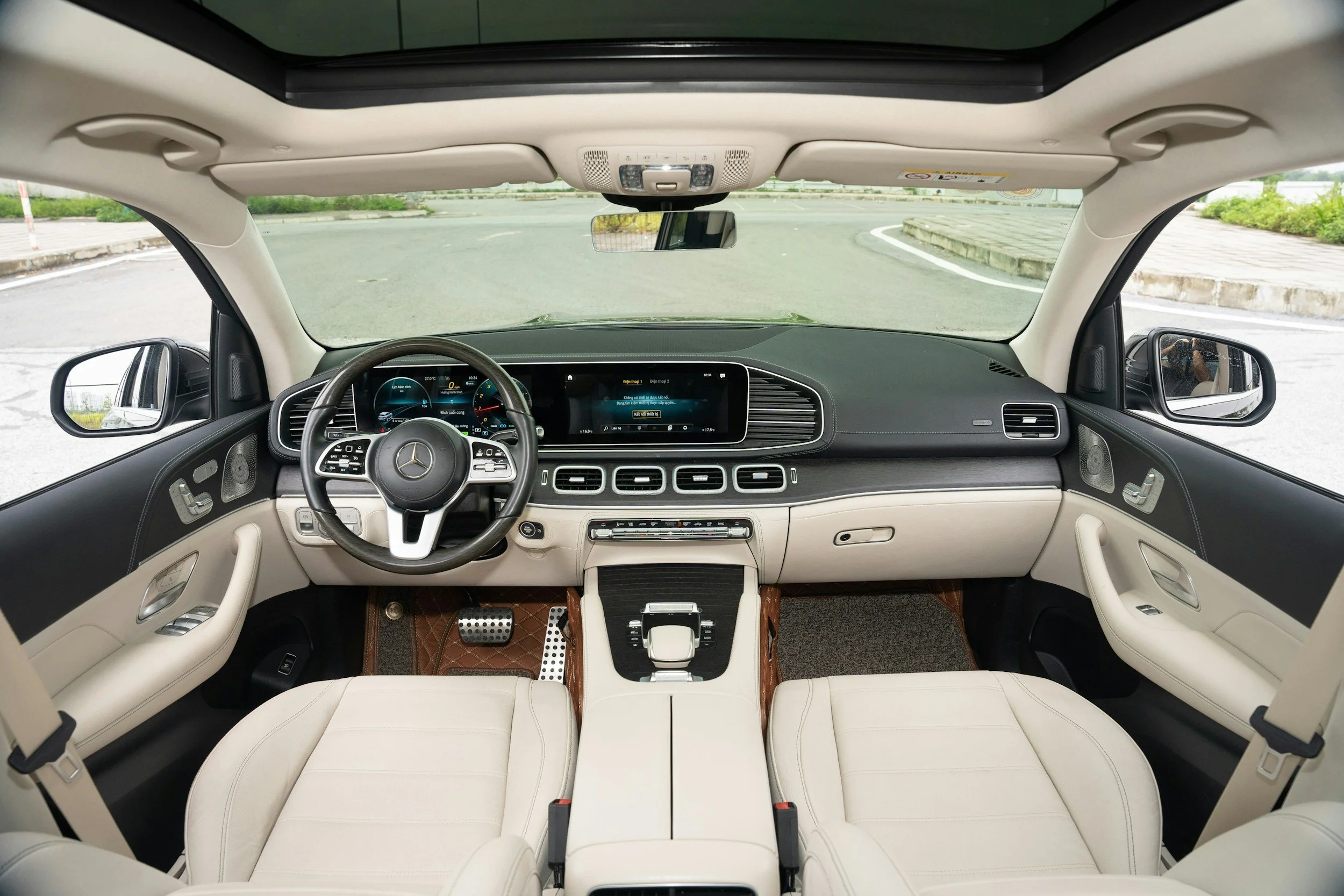 Interior view of a luxury car with a white leather interior, dual front seats, a digital dashboard, and a panoramic sunroof showing a street outside.