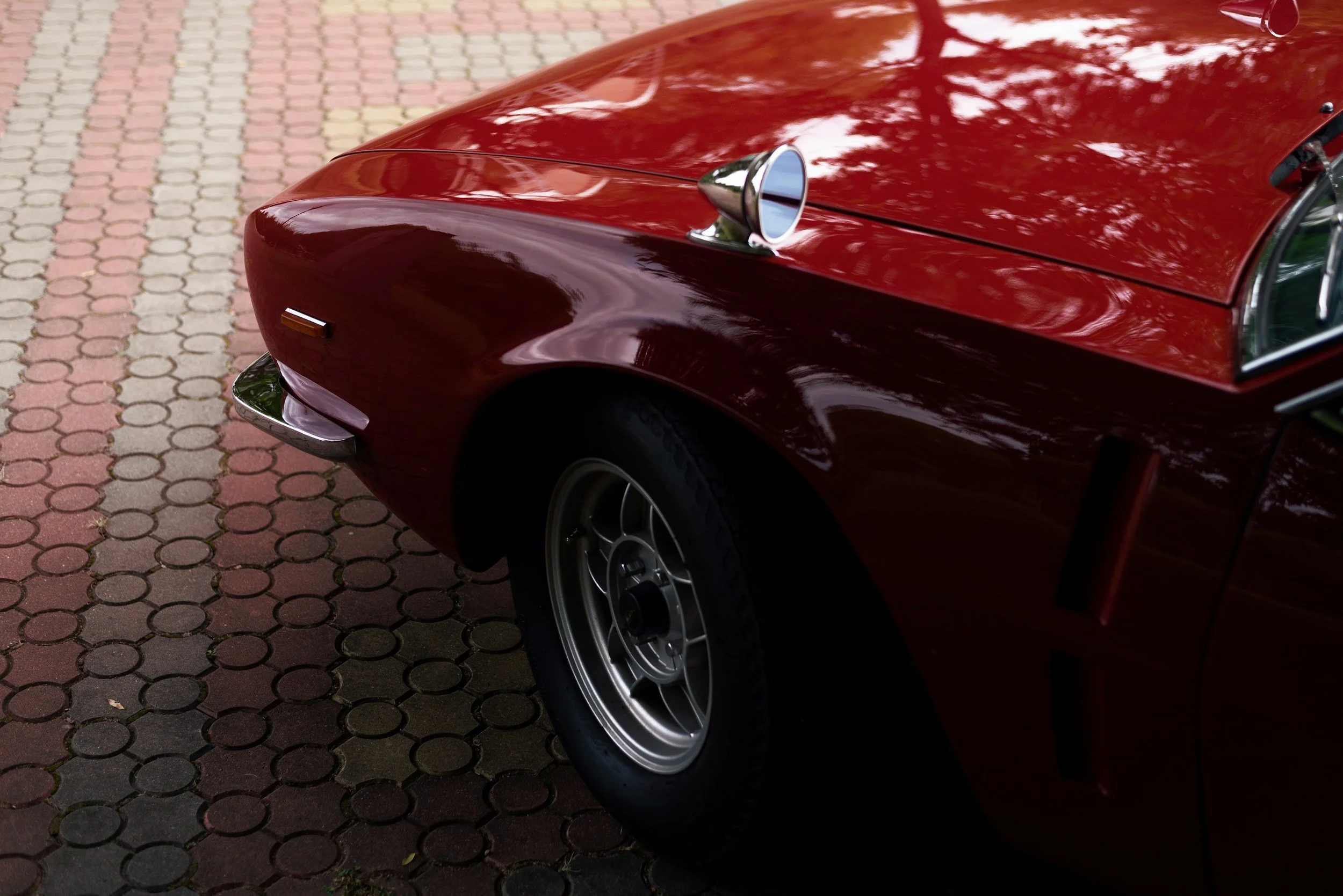 Close-up of a vintage red car's front section, including the hood, window, side mirror, and wheel, parked on a patterned brick pavement.