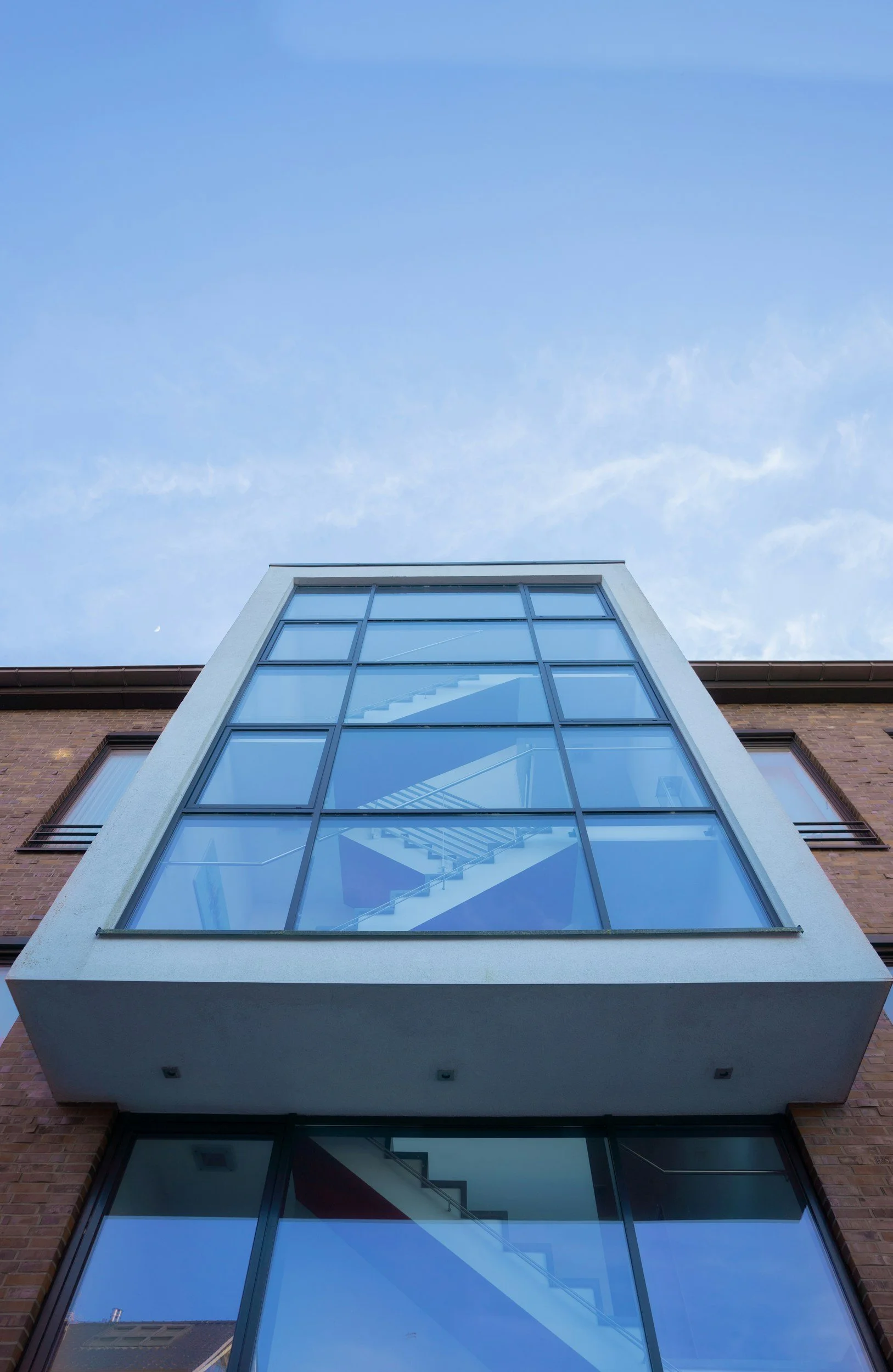 Looking up at a modern building with a large glass window section, brick exterior, and a blue sky with some clouds and a small moon visible above.