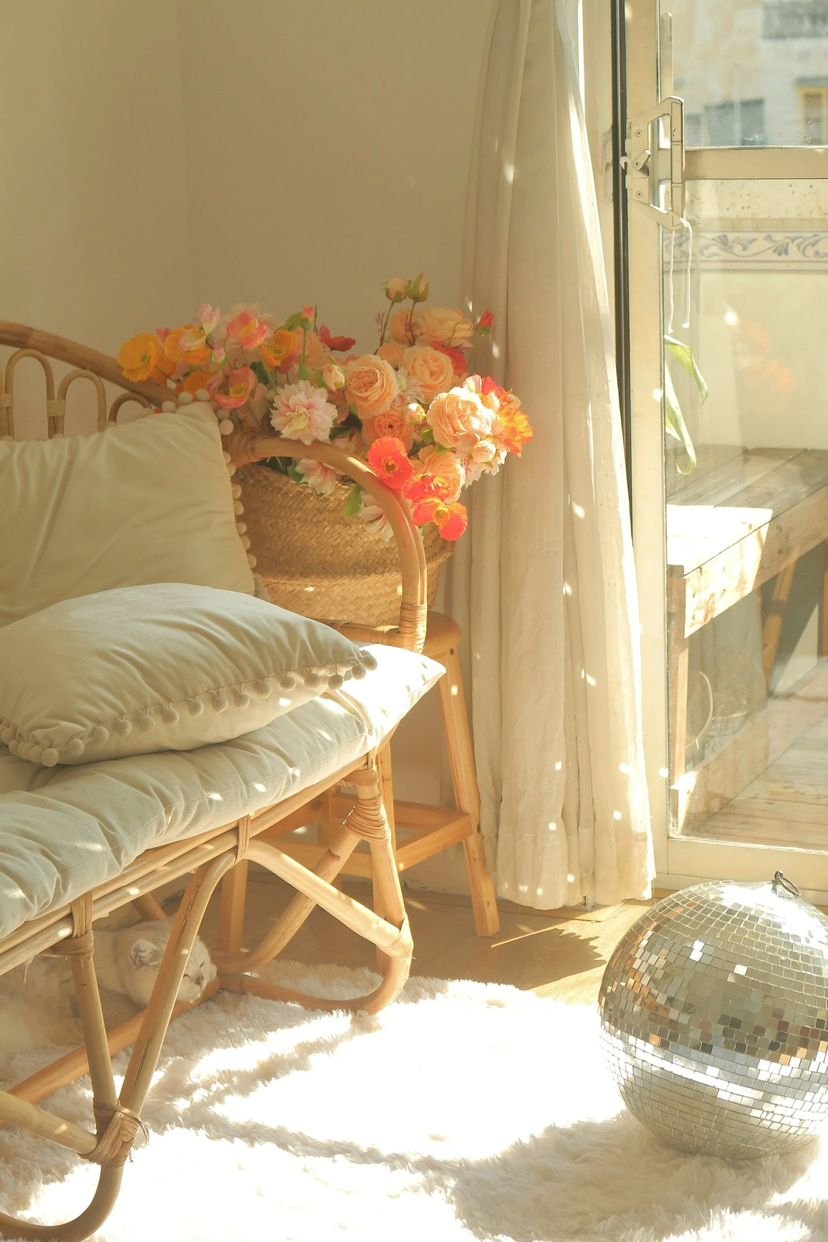 A cozy sunlit corner with a wicker bench draped in white cushions, a vase of colorful flowers, a fluffy white rug, a small white cat underneath the bench, a glass door with curtains, and a mirrored disco ball on the floor.