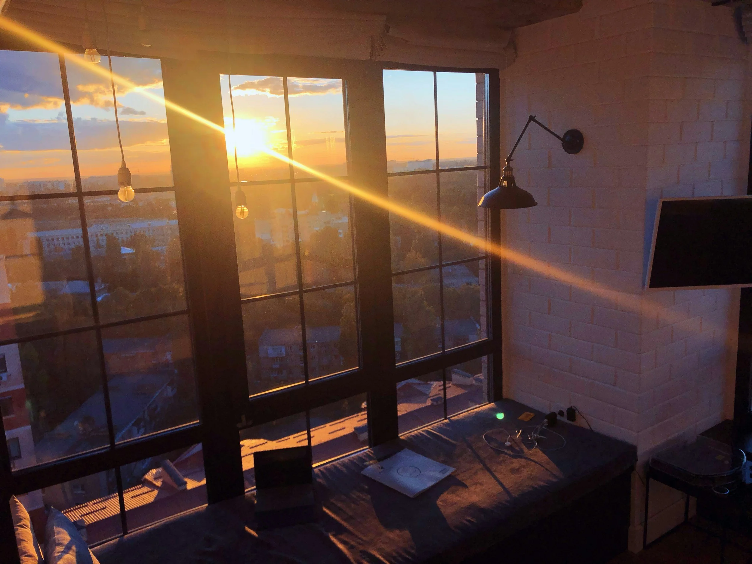 Sunset view through large grid-style windows, sunlight casting warm glow inside the room with a wall-mounted lamp, beams of sunlight creating a pattern on the floor.
