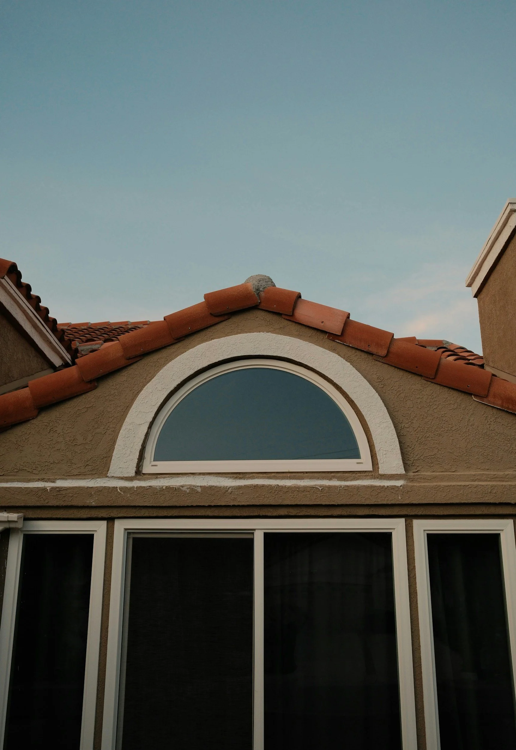 A house exterior with a curved window arched at the top, beige stucco walls, a tiled red roof, and a clear blue sky in the background.