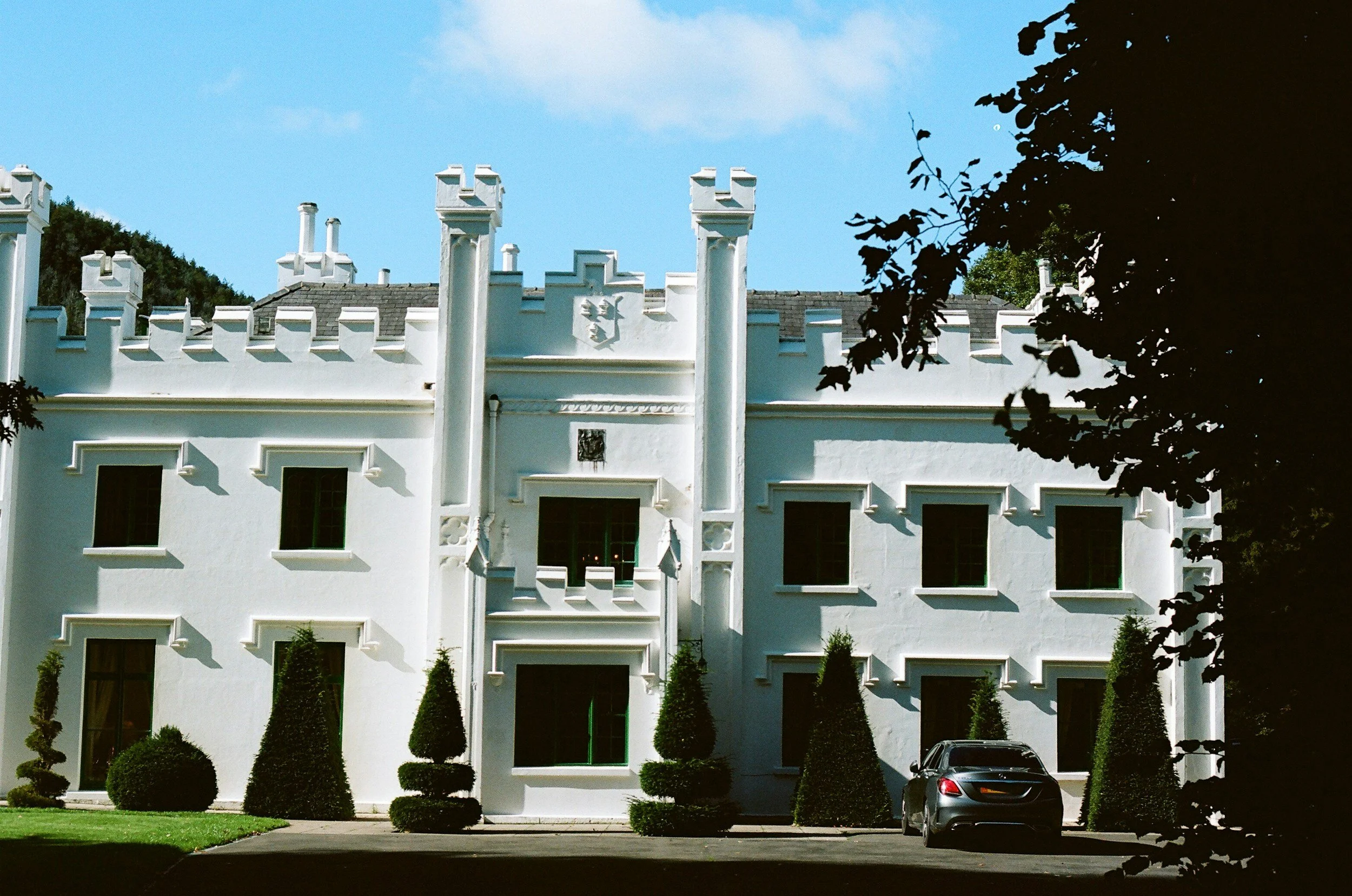 White multi-story building with crenelated roofline and small trees in front, parking lot with a car, and trees casting shadows on the building.