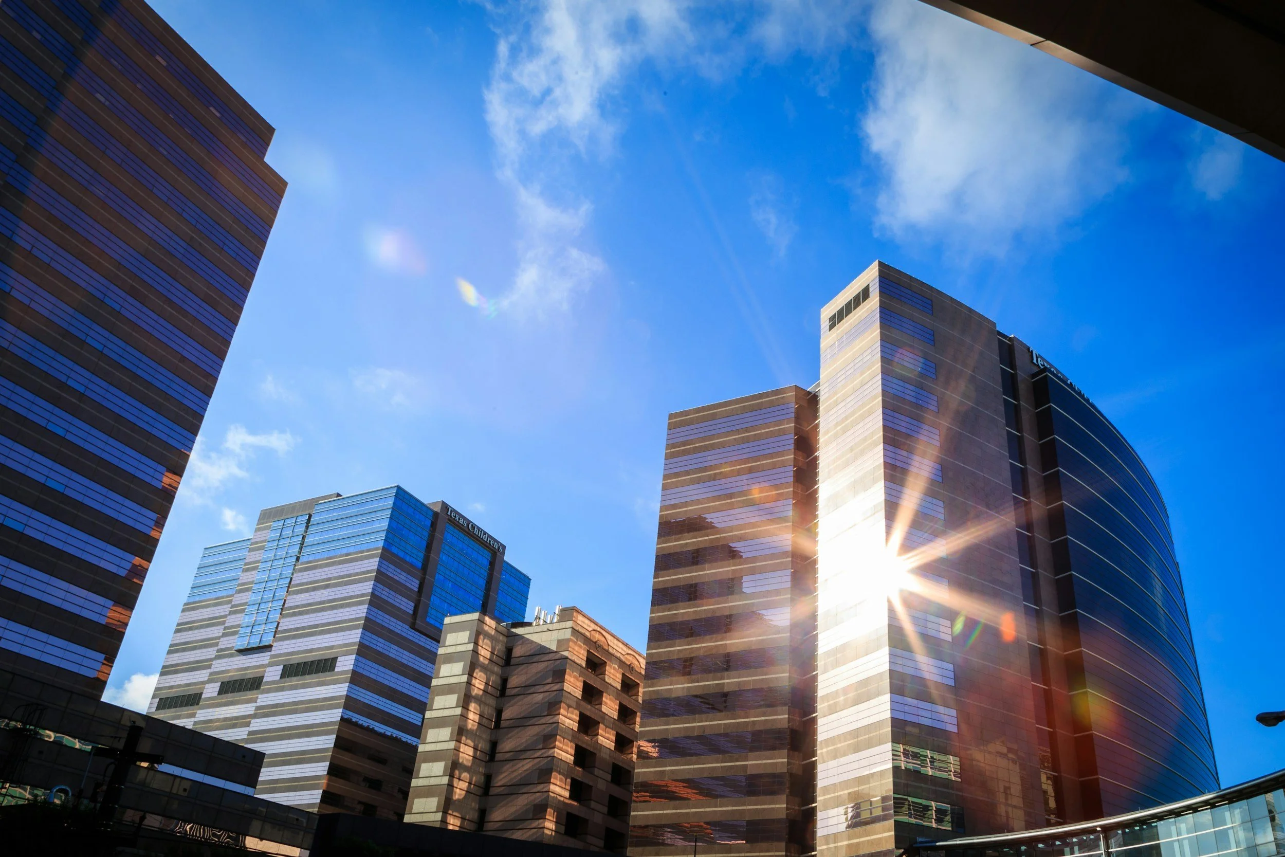 A view of tall modern office buildings with reflective glass windows under a bright blue sky with some clouds. Sunlight creates lens flare in the lower right part of the image.