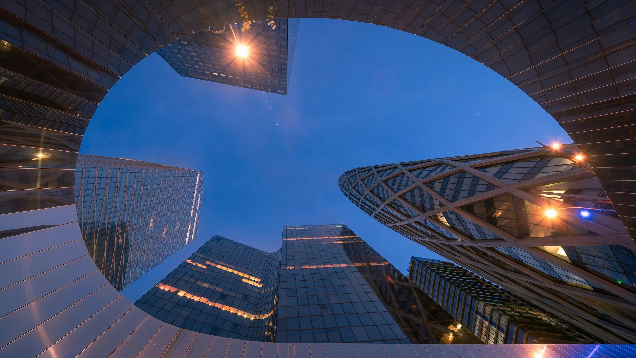 Looking up at tall modern skyscrapers with glass facades in a city at dusk, with the clear blue sky and sunlight reflecting off the buildings.