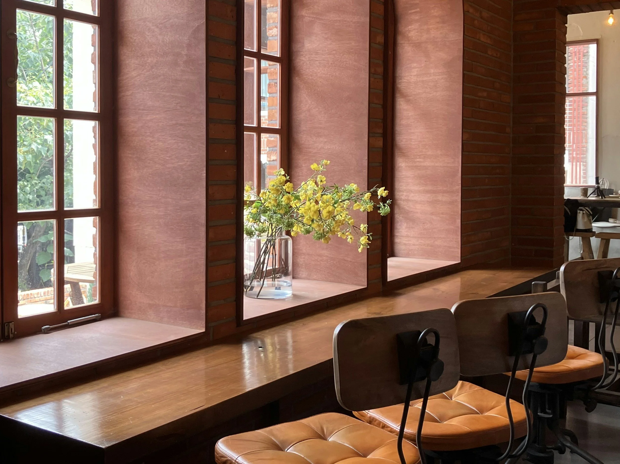 Interior of a room with large windows and a wooden counter, decorated with a vase of yellow flowers, chairs, and brick walls.
